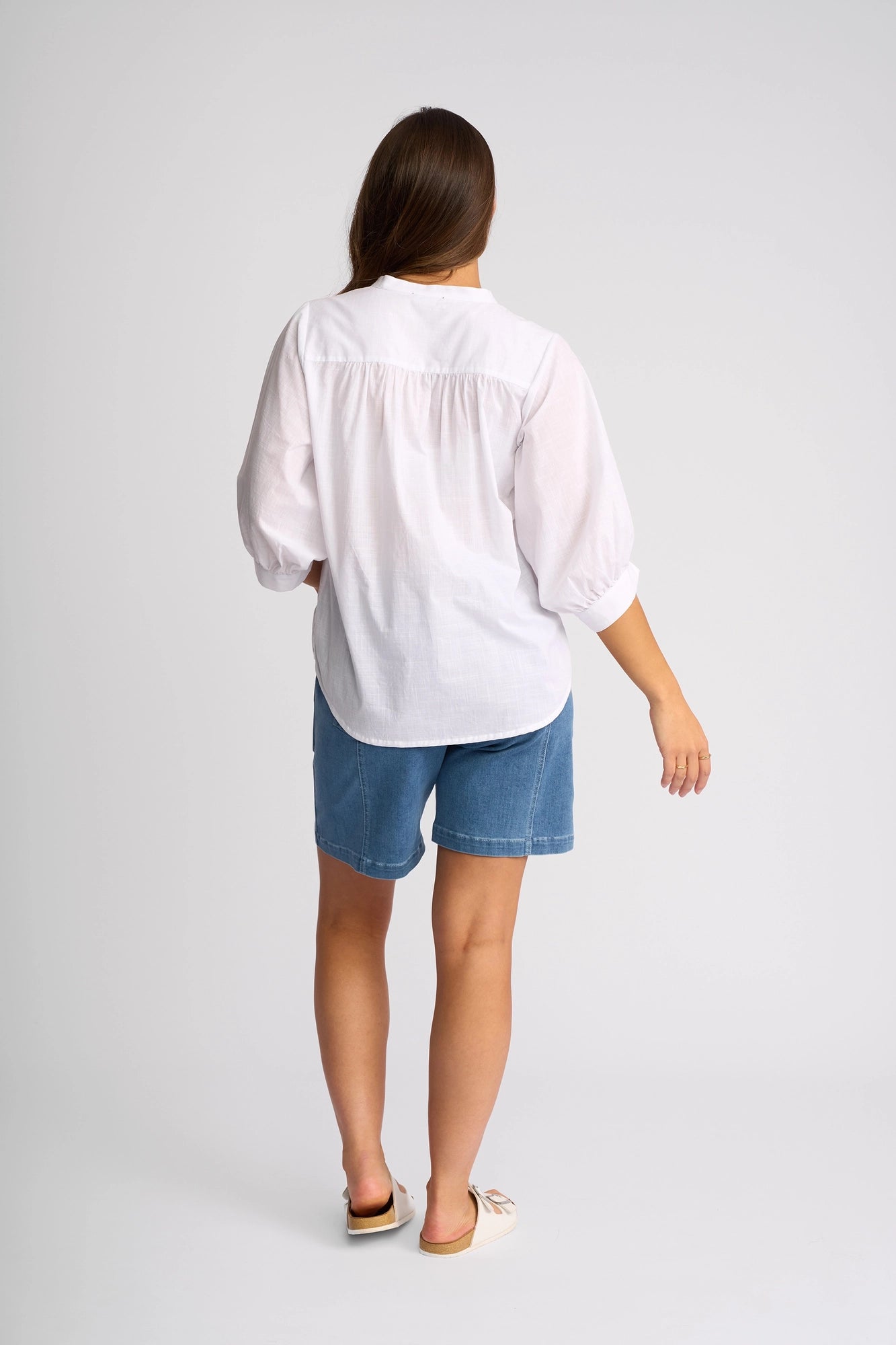 A person stands facing away against a plain light backdrop, wearing the Vassalli Folk Shirt in white—a lightweight cotton shirt with a relaxed silhouette—paired with blue denim shorts and light slip-on shoes.