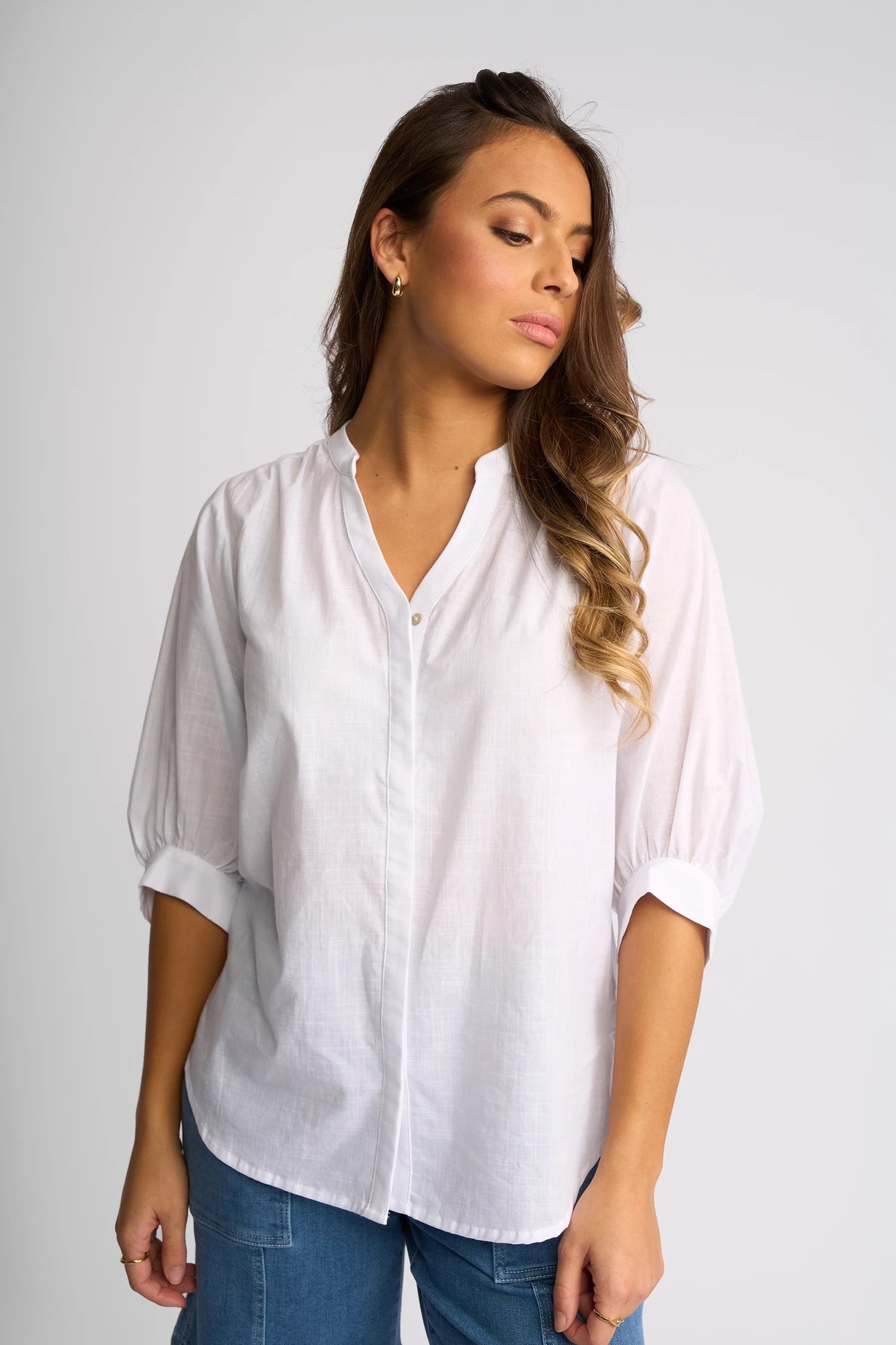 A woman with long wavy hair wears the Vassalli Folk Shirt - White, its relaxed silhouette and lightweight cotton fabric creating an effortless style. She stands against a plain light background, gazing down and to the side with a neutral expression. Blue jeans complete her understated look.