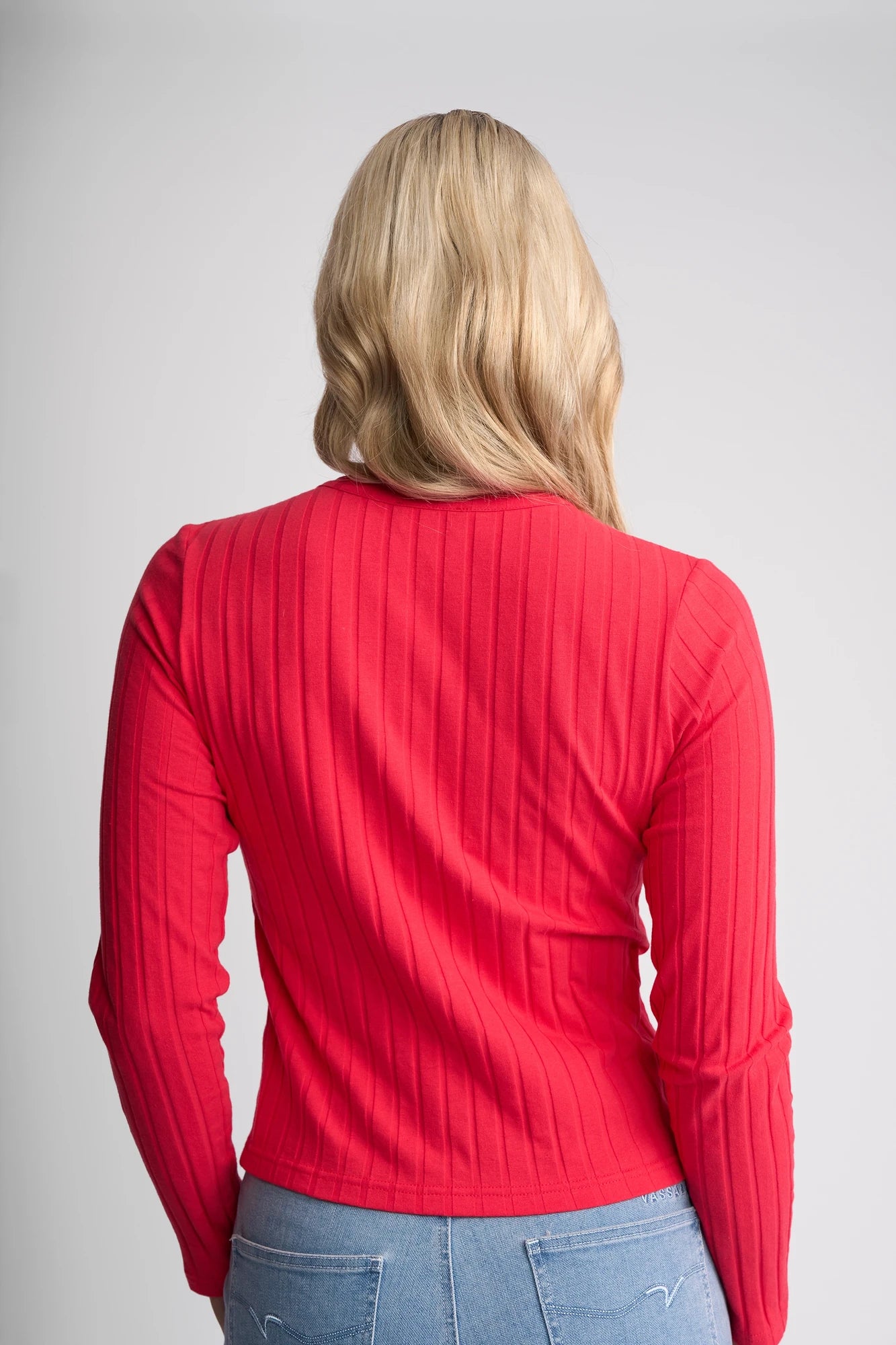 A person with long blonde hair, shown from the back against a plain light gray background, is wearing the Vassalli Suri Cardi in Cherry—a red ribbed stretch cotton long-sleeve top—paired with light blue jeans.