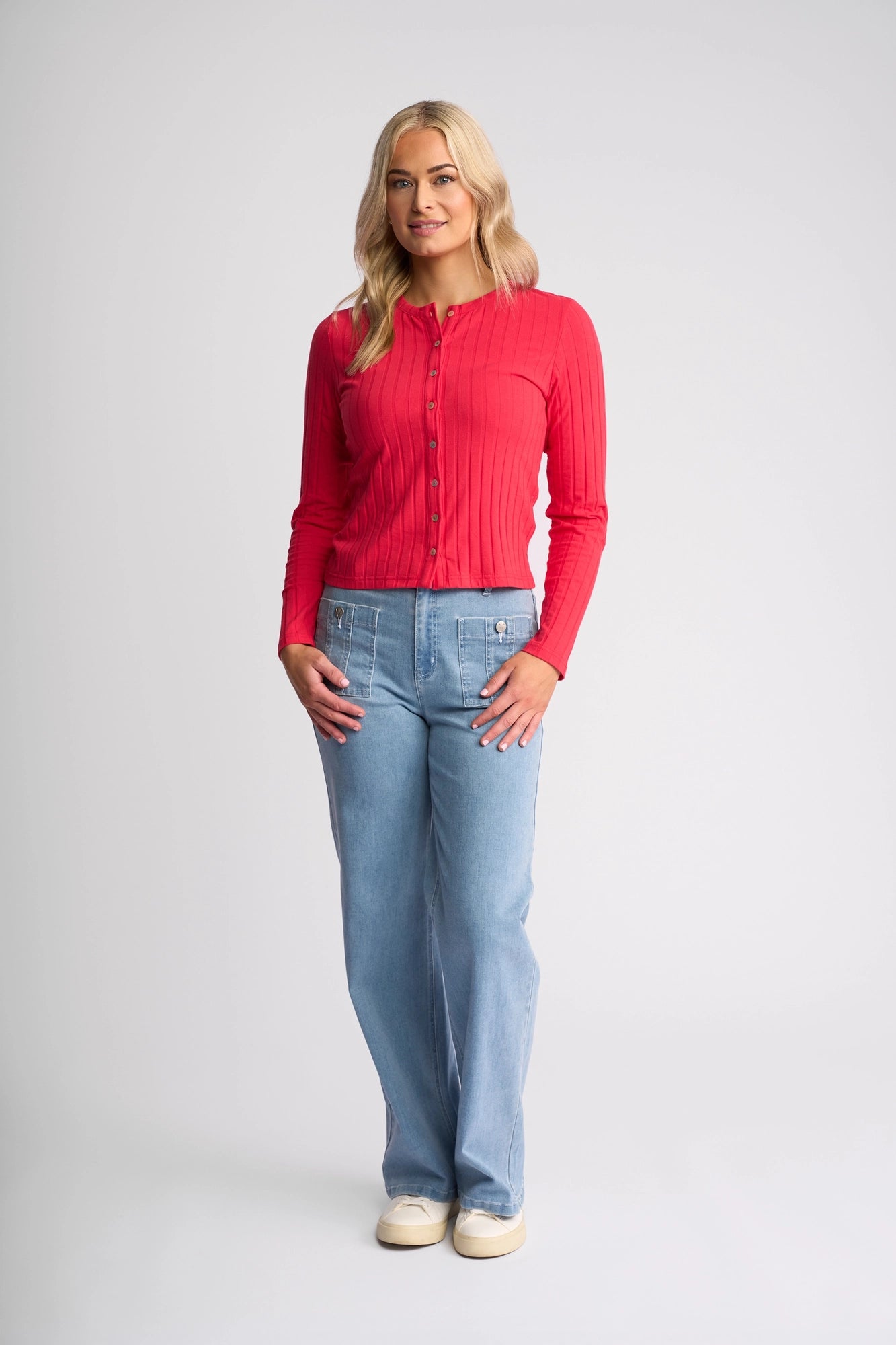 A woman with long blonde hair and a neutral expression stands facing forward against a plain light background. She is wearing the Vassalli Suri Cardi in Cherry—a bright red, slim-fitting cardigan with a classic crew neckline—paired with light blue jeans and white shoes.