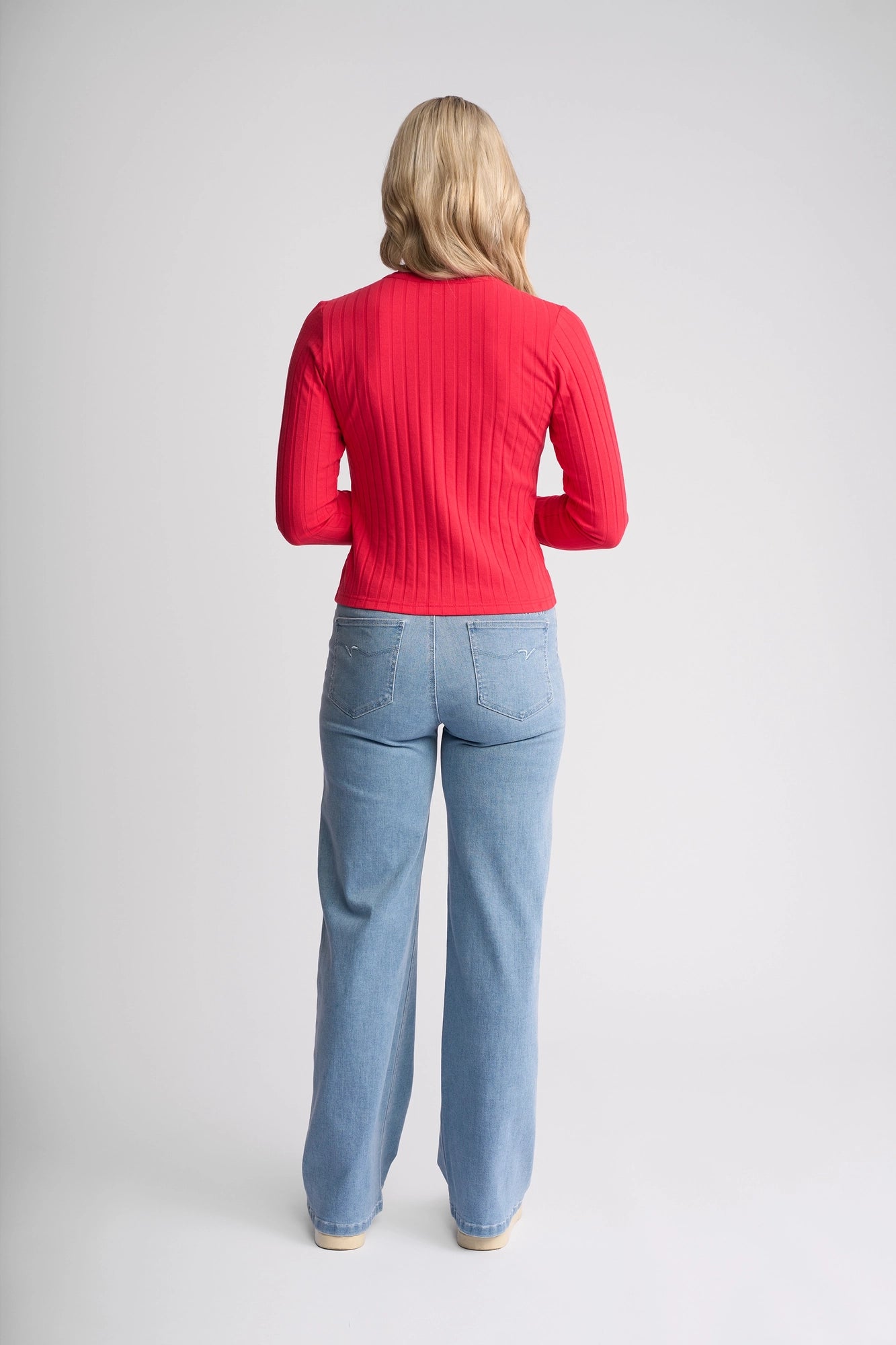 A person with long blonde hair, wearing the Vassalli Suri Cardi in Cherry—a red long-sleeve ribbed stretch cotton top with a crew neckline—and light blue jeans, stands facing away from the camera against a plain white background.