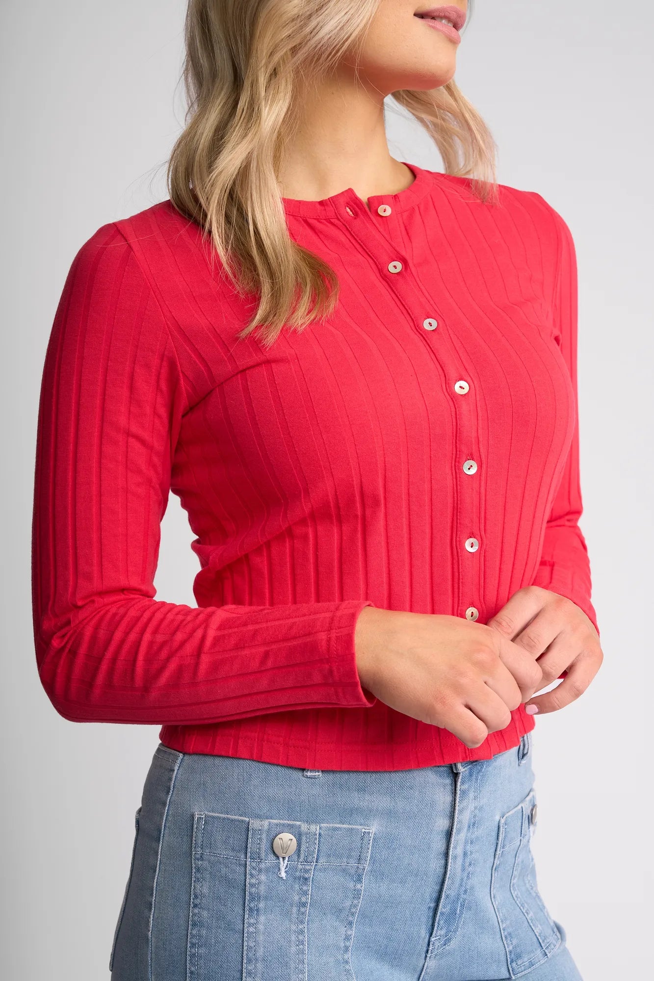 A woman wearing the Vassalli Suri Cardi in Cherry—a slim-fitting, bright red ribbed stretch cotton button-up top with a crew neckline—and a light blue denim skirt with visible pockets, standing against a plain background.