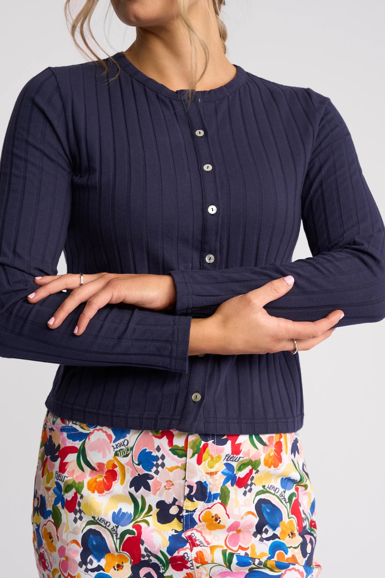 A woman with crossed arms wears the Vassalli Suri Cardi in navy, featuring a ribbed button-up design, paired with colorful floral print pants. Her face is partially out of frame, and her hair is tied back.