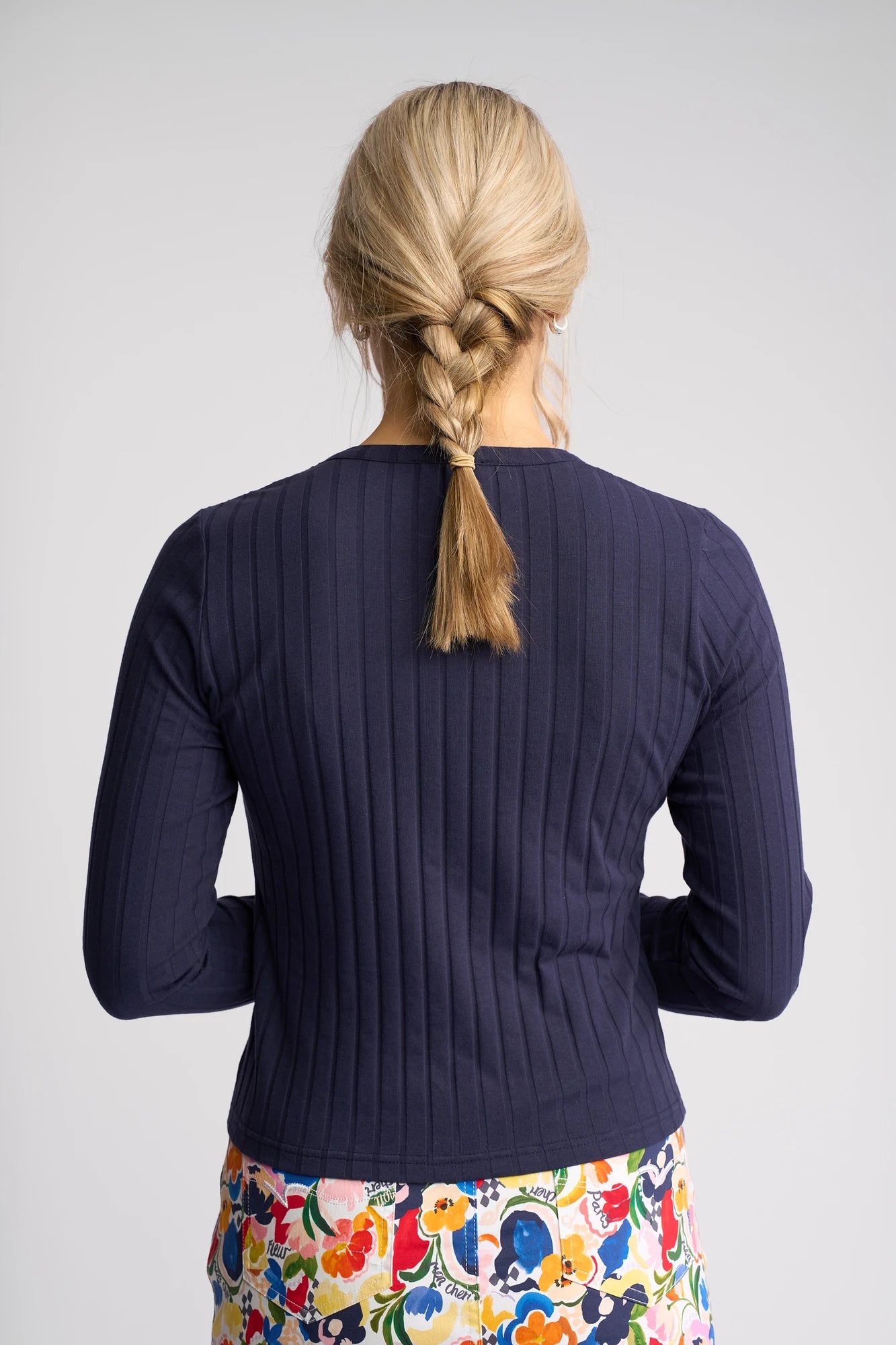 A woman with blonde hair in a braid, wearing the Vassalli Suri Cardi in Navy paired with a colorful floral skirt, stands facing away from the camera against a plain background.