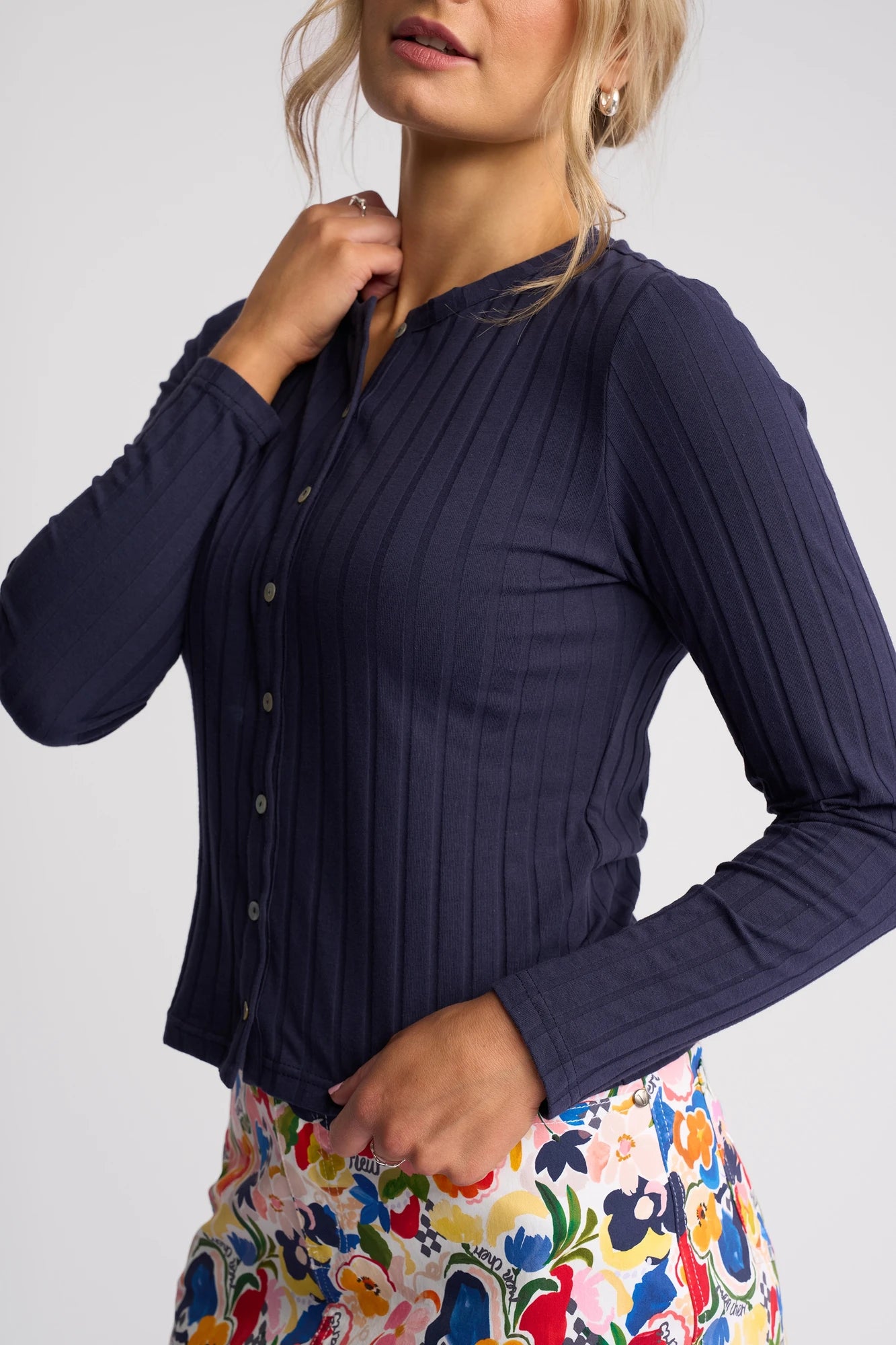 A woman stands against a plain light background, wearing the Vassalli Suri Cardi in navy—a slim-fitting stretch cotton cardigan—paired with colorful floral pants. Her head is slightly turned and one hand gently touches her neck.