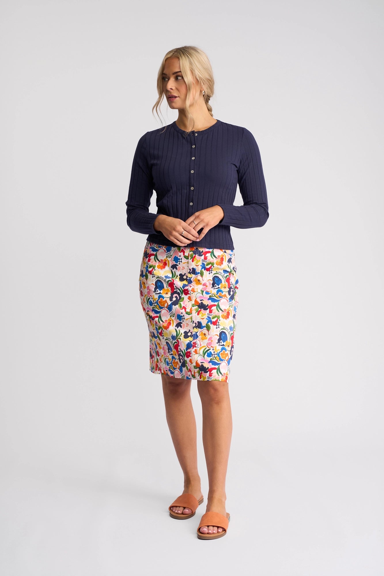 A woman stands in a studio wearing the Vassalli Suri Cardi in Navy layered over a colorful floral skirt and tan sandals, her hands clasped in front as she looks to the side.