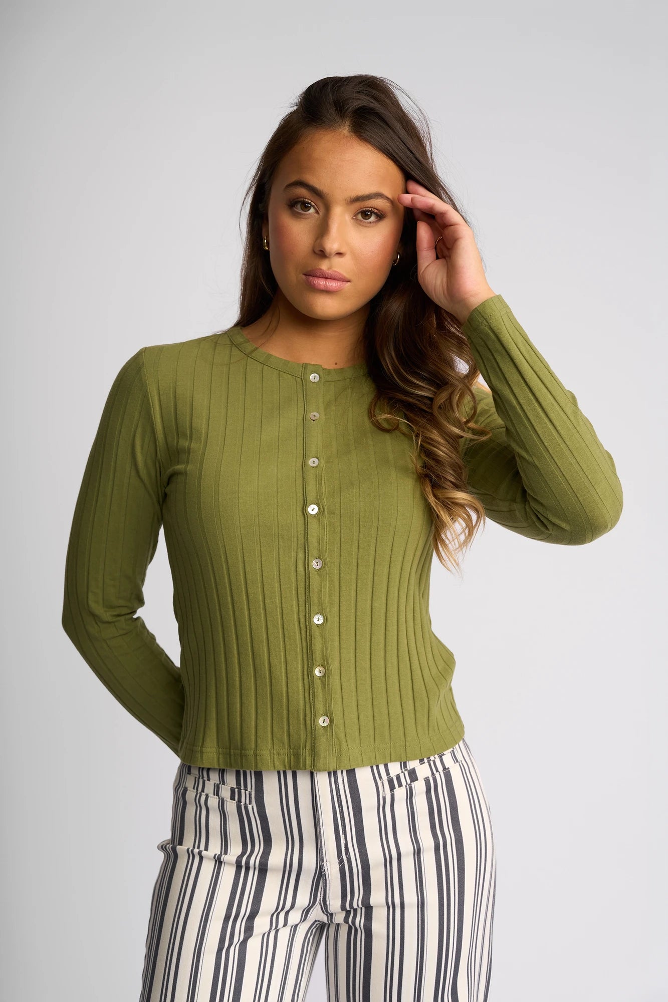 Wearing the Vassalli Suri Cardi in olive, a woman stands against a plain background in white pants with black vertical stripes, gazing forward with one hand touching her hair.