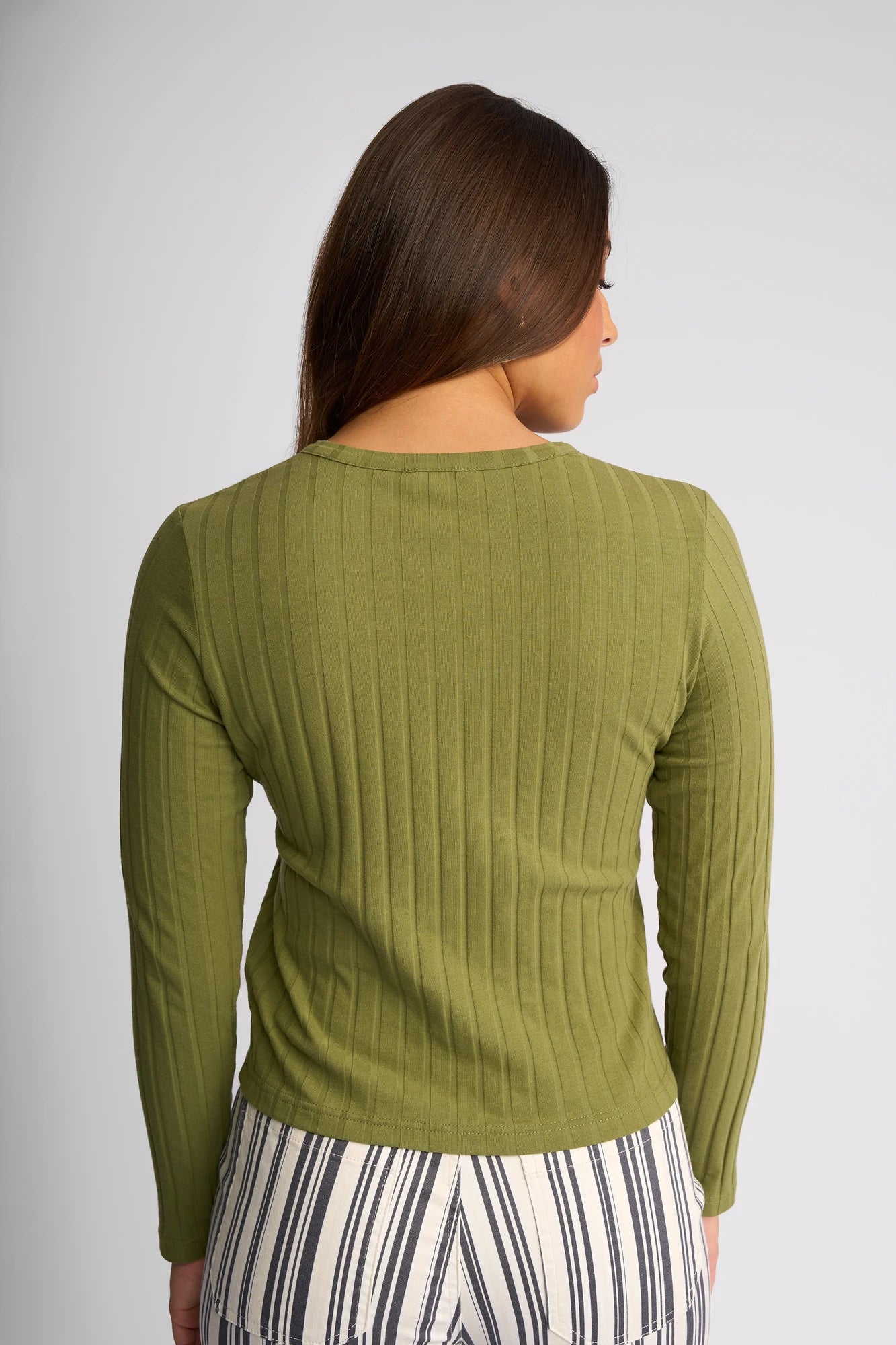 A woman with long brown hair, shown from the back, wears the Vassalli Suri Cardi in olive and high-waisted black and white vertically striped pants against a plain light background.