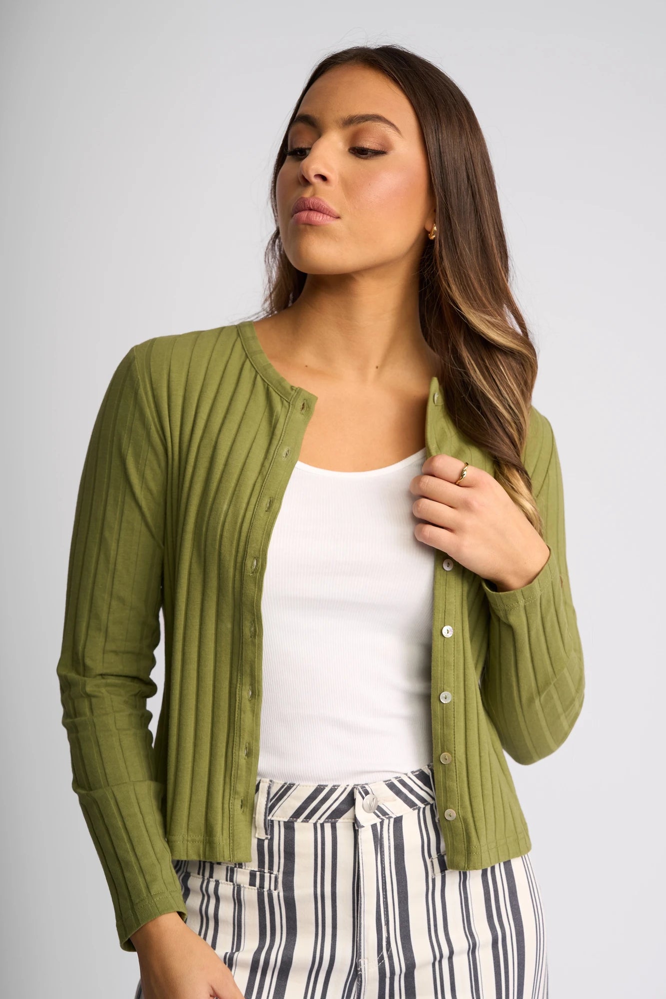 Wearing the Vassalli Suri Cardi in Olive over a white tank top and striped pants, a woman looks to the side with her hand touching the cardigan’s buttons.