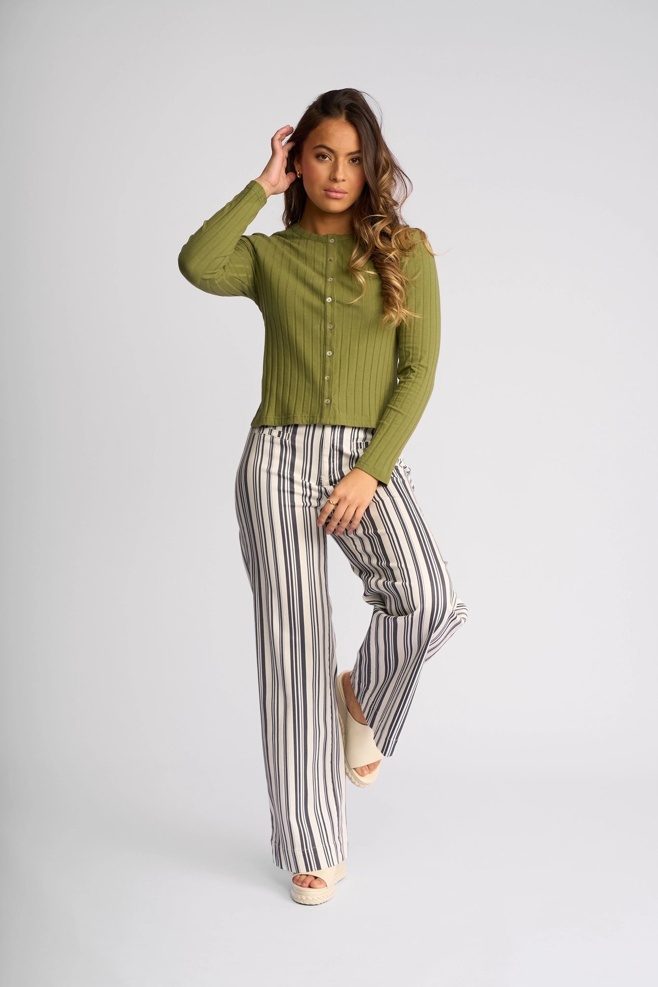 A woman poses in the Vassalli Suri Cardi in olive, styled with high-waisted, wide-leg white pants featuring vertical black stripes and wedge sandals, set against a plain white background.