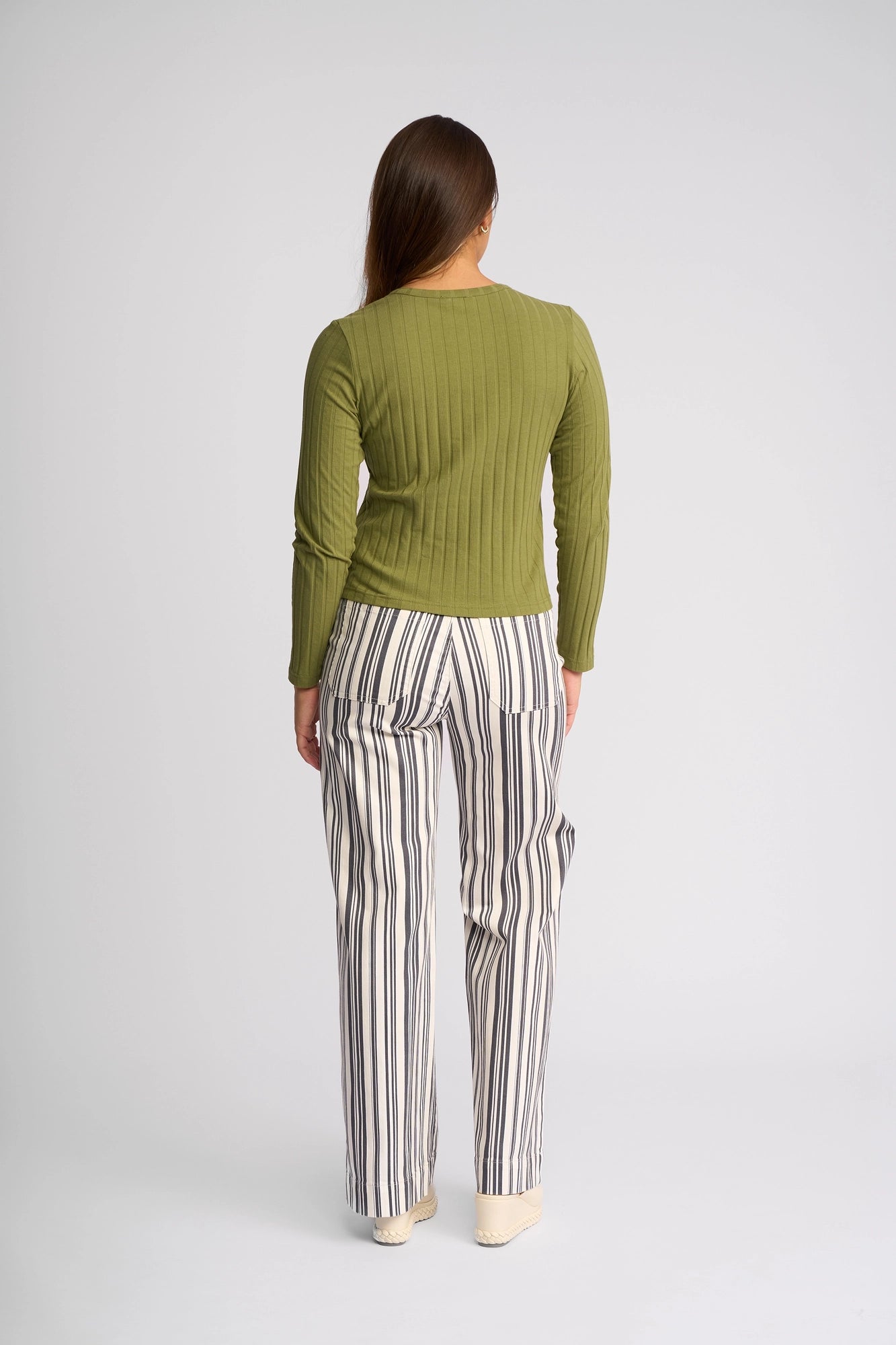 A person with long brown hair stands facing away against a plain light background, wearing the Vassalli Suri Cardi in olive and white pants with vertical black stripes.
