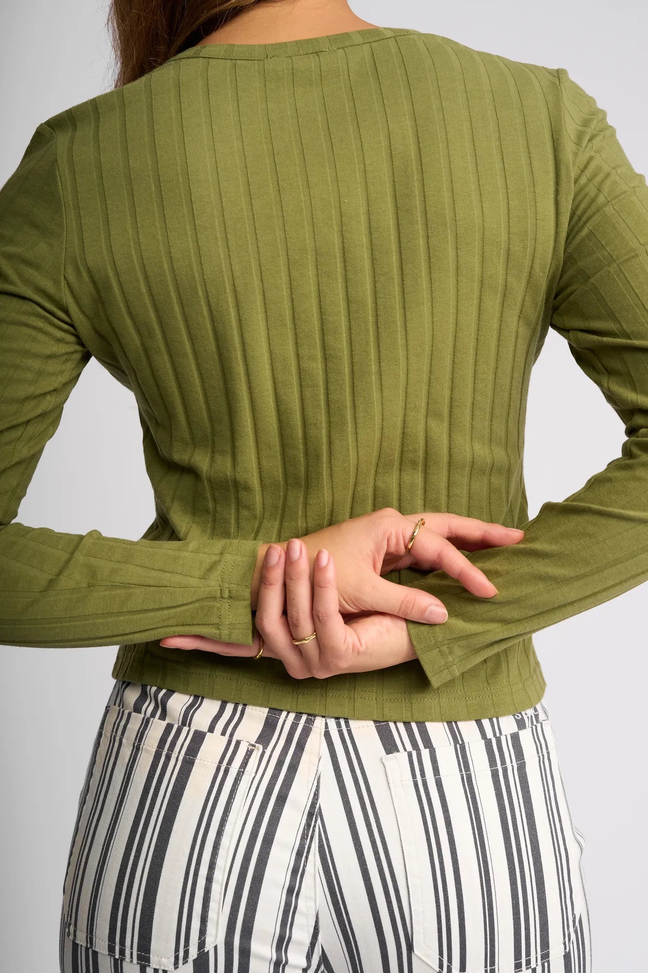 Wearing the Vassalli Suri Cardi in olive, a person stands with their back to the camera, one hand behind their back holding the other wrist, paired with striped pants.
