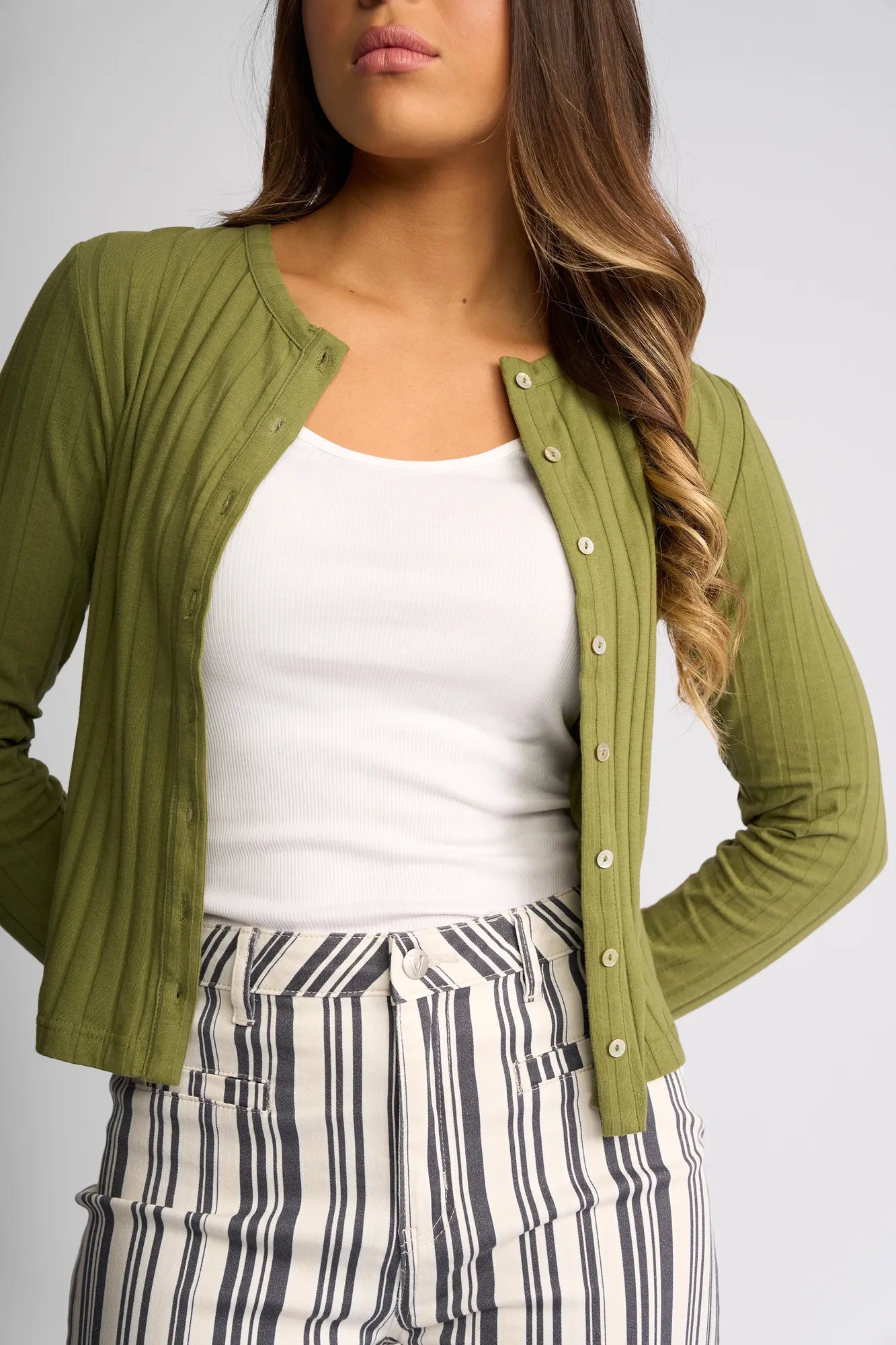 A woman wearing the Vassalli Suri Cardi in olive over a white tank top and high-waisted, black-and-white striped pants stands with her hands on her hips.