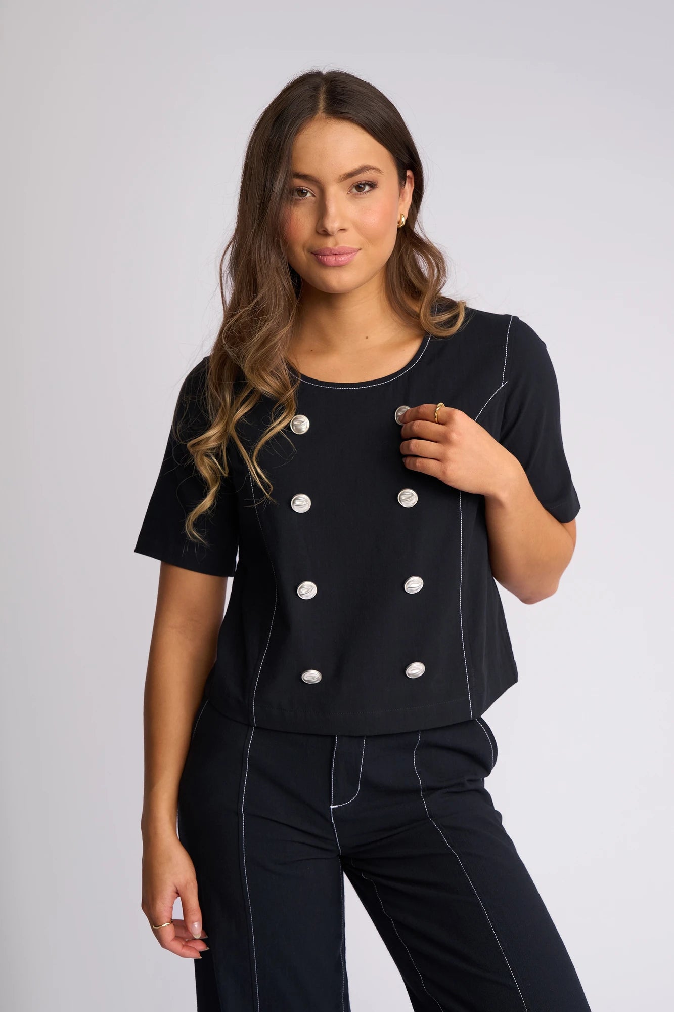 A woman with long wavy hair wears the Vassalli Oslo Top in Ink—a black, short-sleeve shirt featuring white stitching and two rows of silver buttons—paired with matching black pants, standing against a plain background.