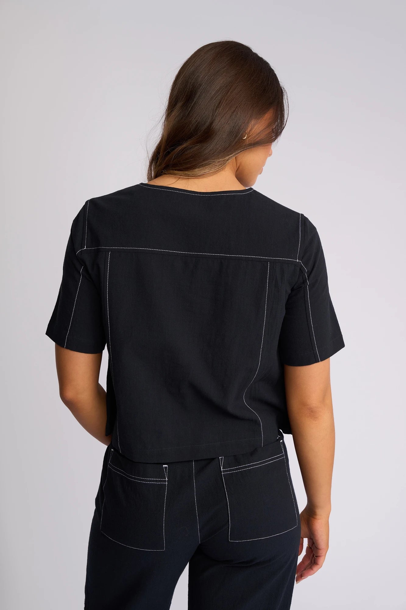 A person with long brown hair wearing a black short-sleeved Vassalli Oslo Top - Ink, crafted from 100% cotton, and black pants with white stitching stands facing away from the camera.
