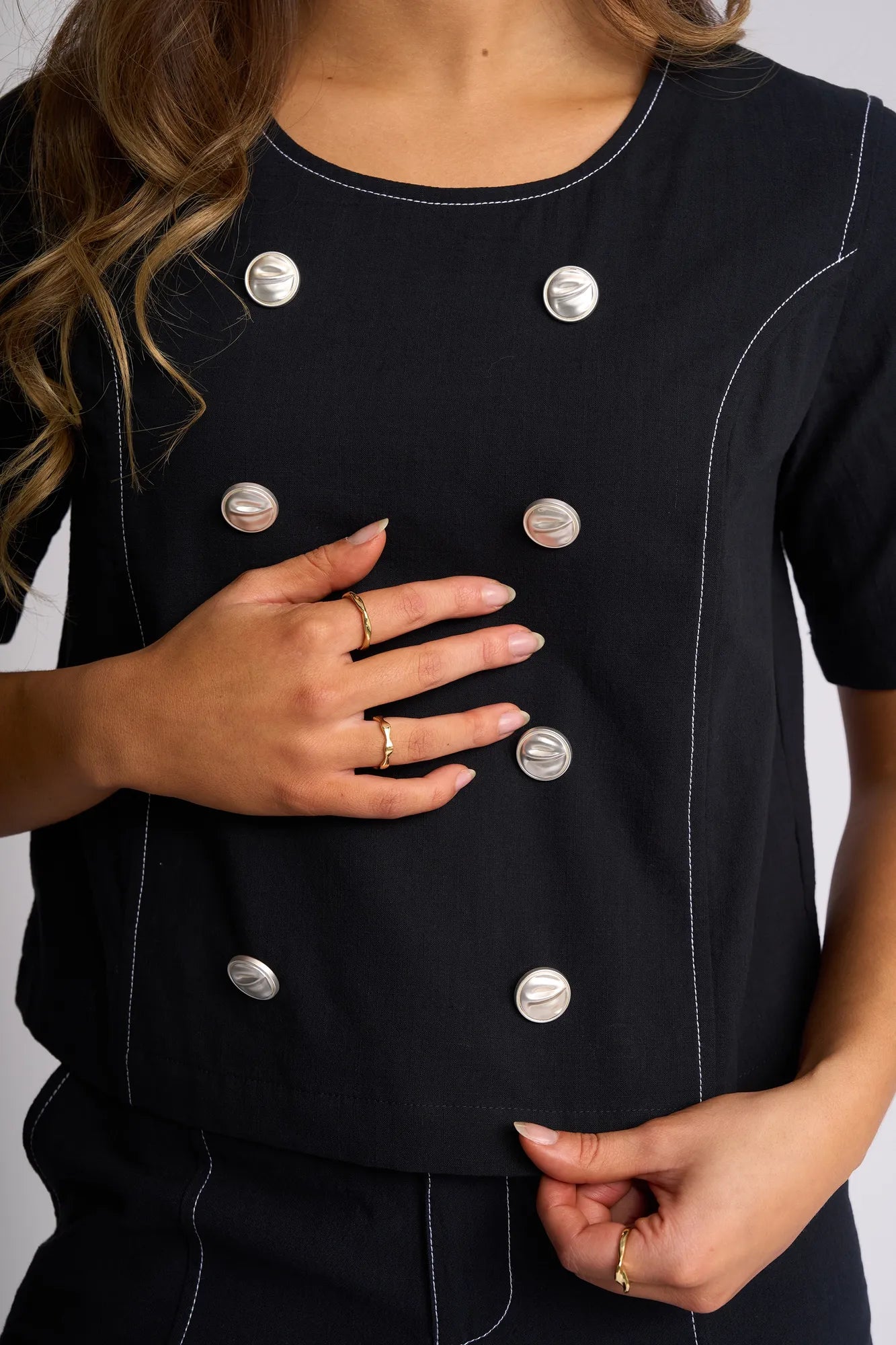 A person wearing a Vassalli Oslo Top - Ink, featuring short sleeves, double-breasted design with white stitching and two rows of large silver buttons, rests their hand on their stomach, displaying several gold rings on their fingers.
