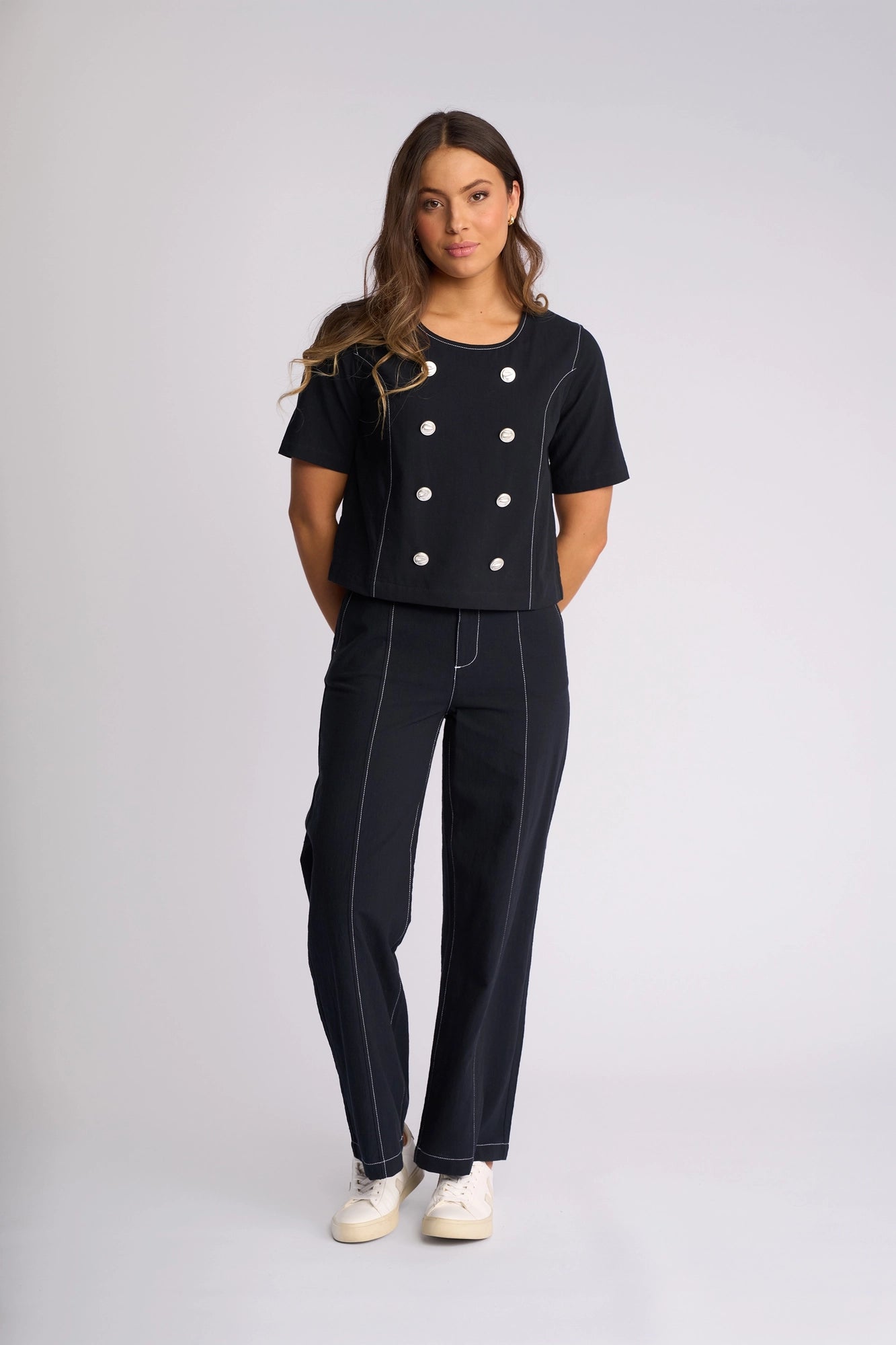 A woman stands against a light background, wearing the Vassalli Oslo Top in Ink—a 100% cotton, black double-breasted short-sleeve top with white buttons—paired with matching black pants featuring white stitching and finished with white sneakers.