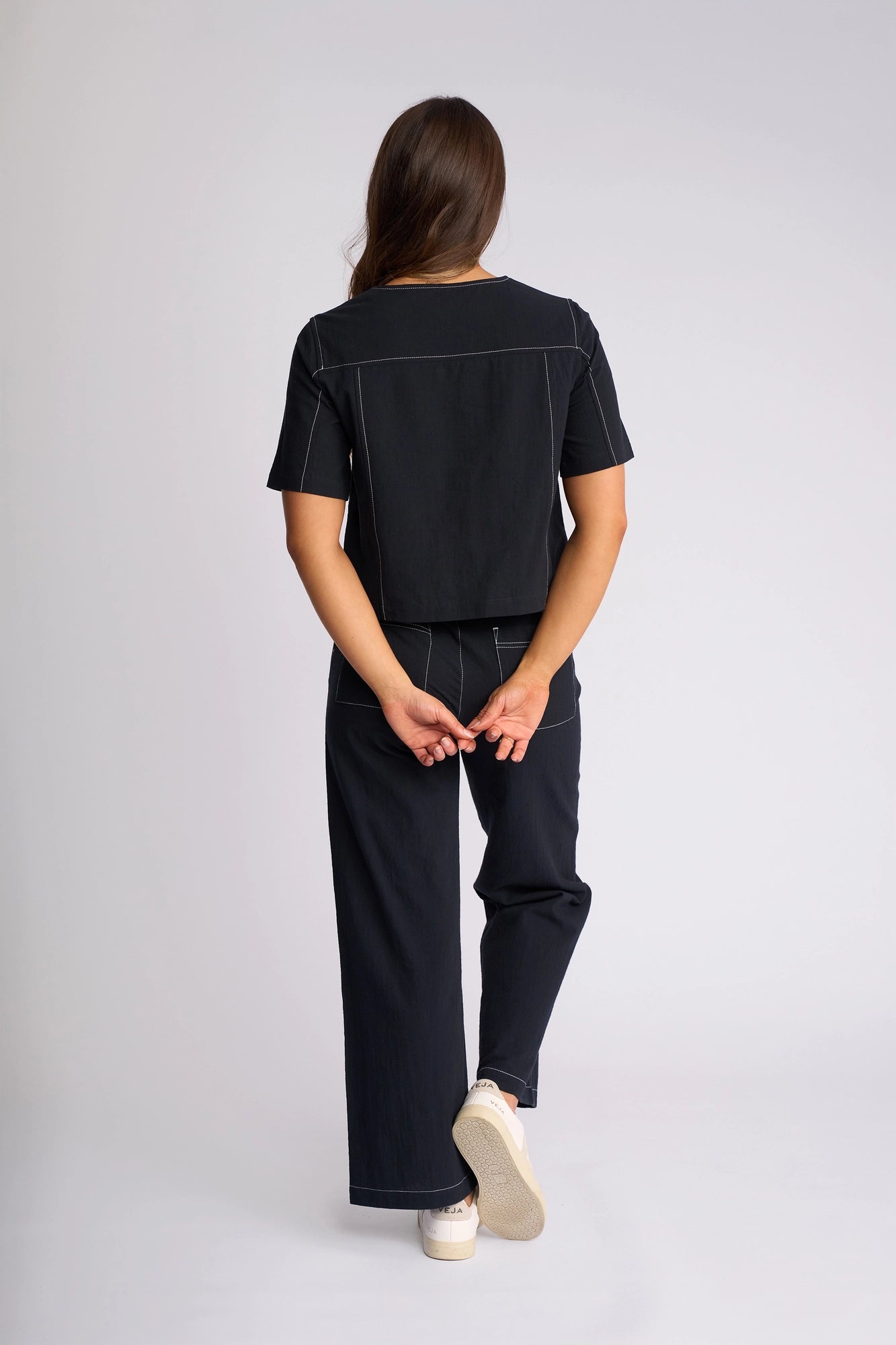 A person stands facing away, hands clasped behind their back. They wear a Vassalli Oslo Top in Ink—a black short-sleeve shirt—paired with black wide-leg pants featuring white stitching, both made from 100% cotton, and white sneakers. The background is plain light gray.