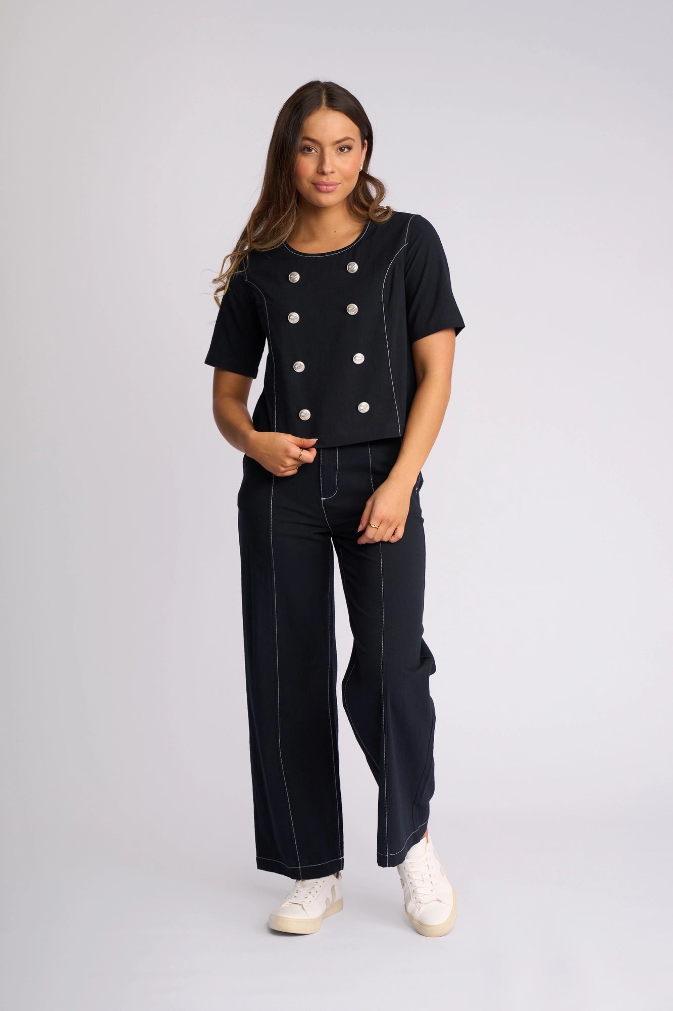A woman stands against a plain background, wearing the Vassalli Oslo Top in Ink—a double-breasted black two-piece outfit featuring white buttons and white stitching, crafted from 100% cotton—and paired with white sneakers.