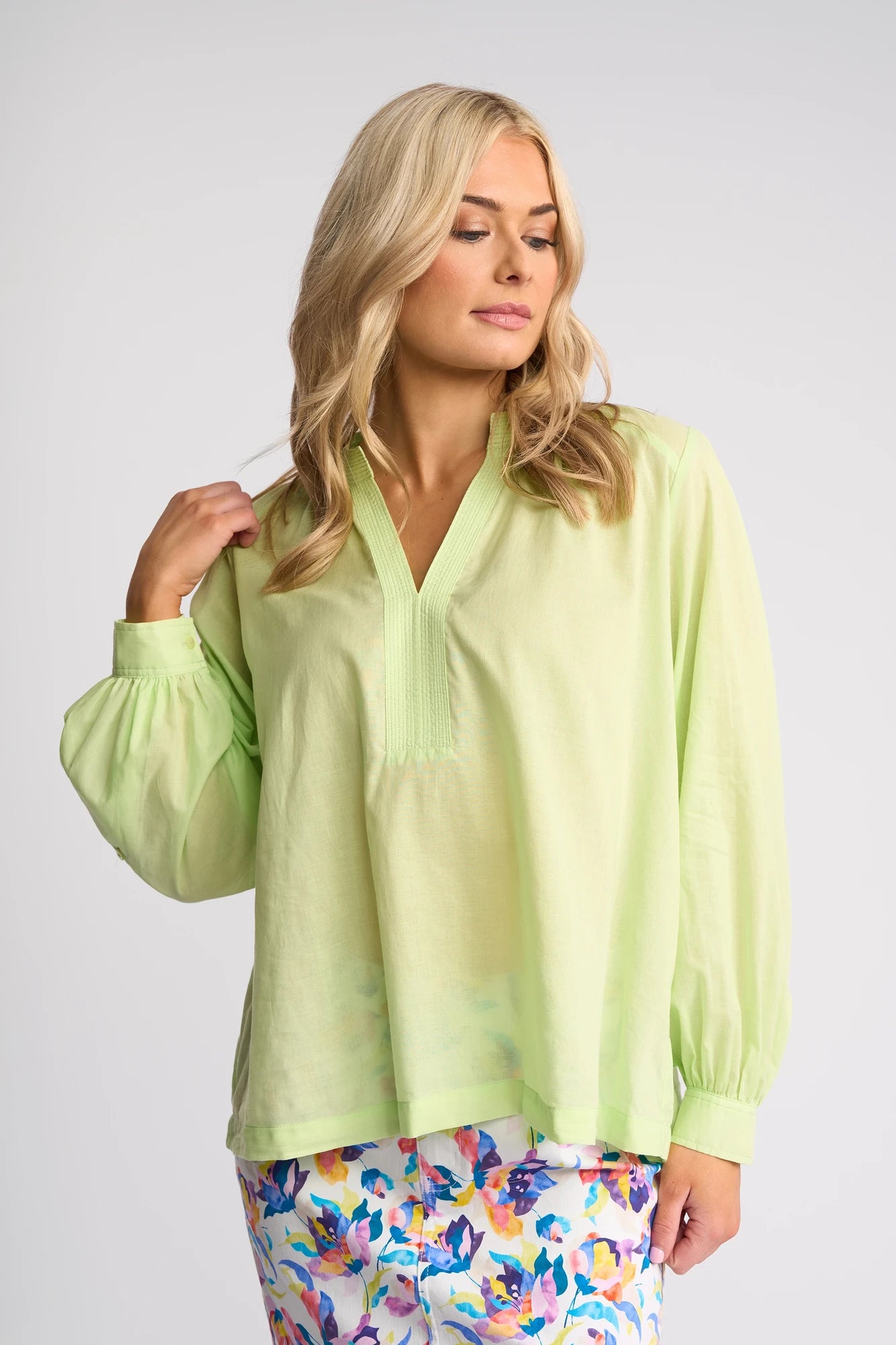 A woman with long blonde hair wears the Vassalli Lily Shirt in Citrus—a light green, lightweight, long-sleeved shirt—and colorful floral pants, standing against a plain light background.