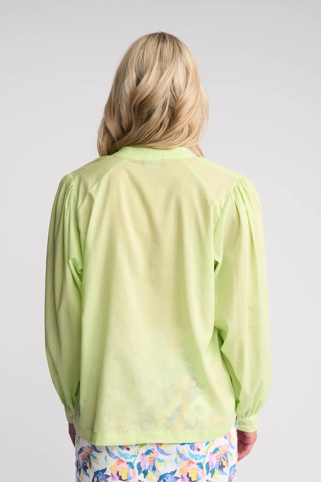 A person with long blonde hair is seen from the back, wearing a light green Vassalli Lily Shirt - Citrus and floral-patterned pants, standing against a plain light gray background.