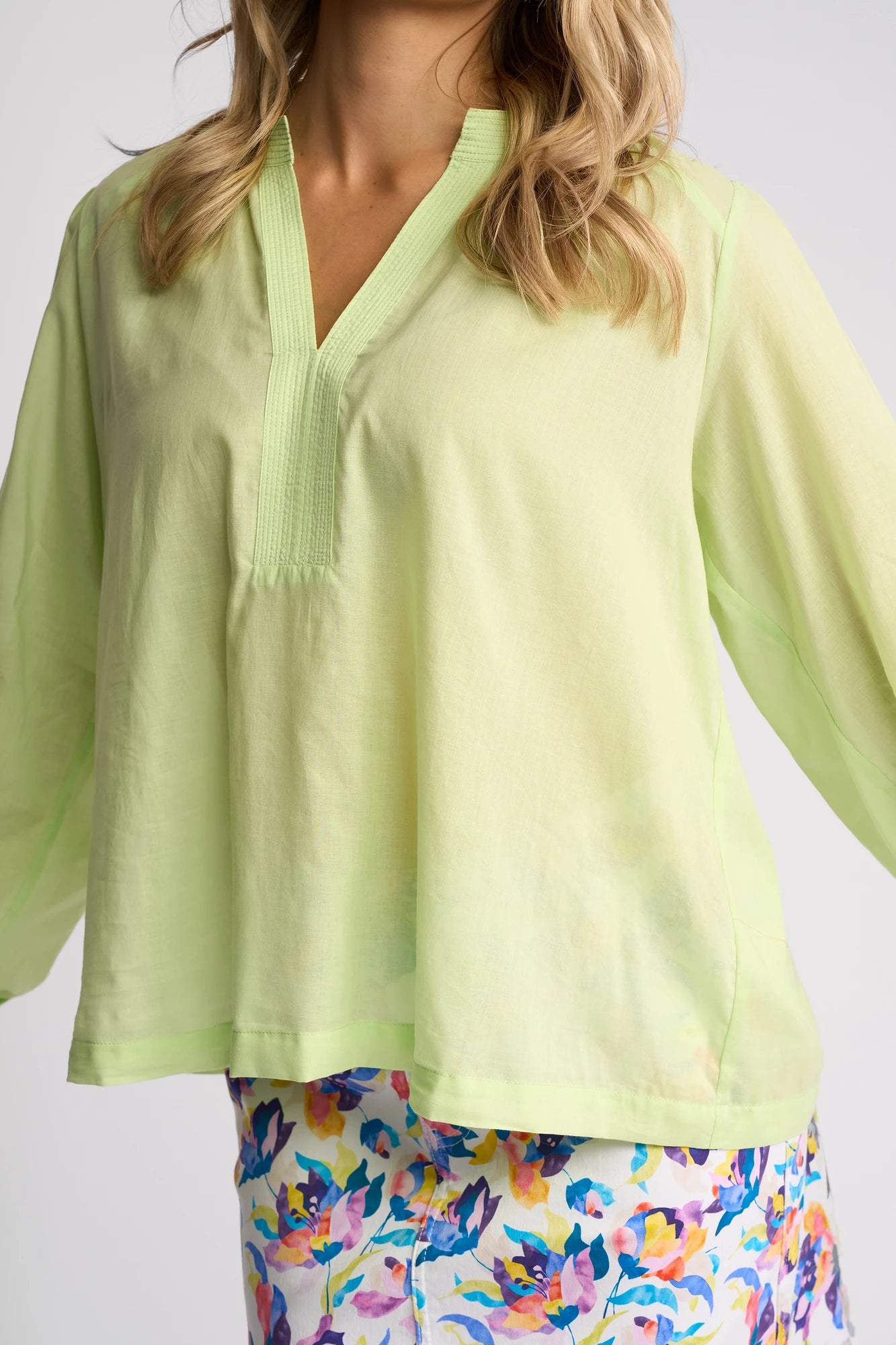 A woman is wearing a loose, citrus green Vassalli Lily Shirt with a V-neck and quarter sleeves, paired with colorful, floral patterned shorts. Only her upper body is visible.