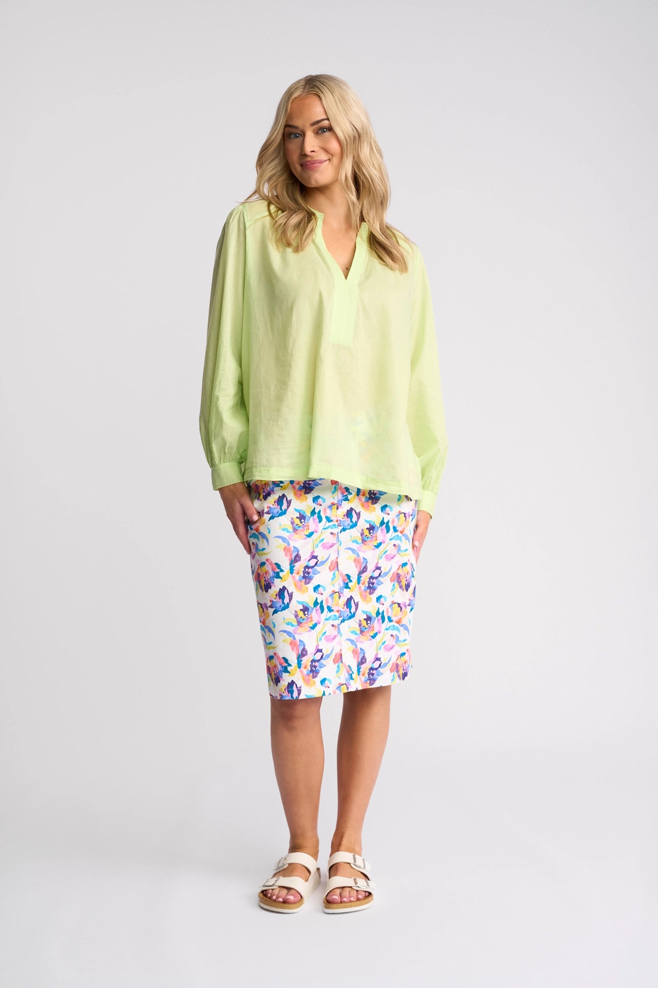 A woman stands smiling in a light green Lily Shirt - Citrus by Vassalli, paired with a knee-length white skirt featuring colorful floral patterns and white sandals against a plain light background.