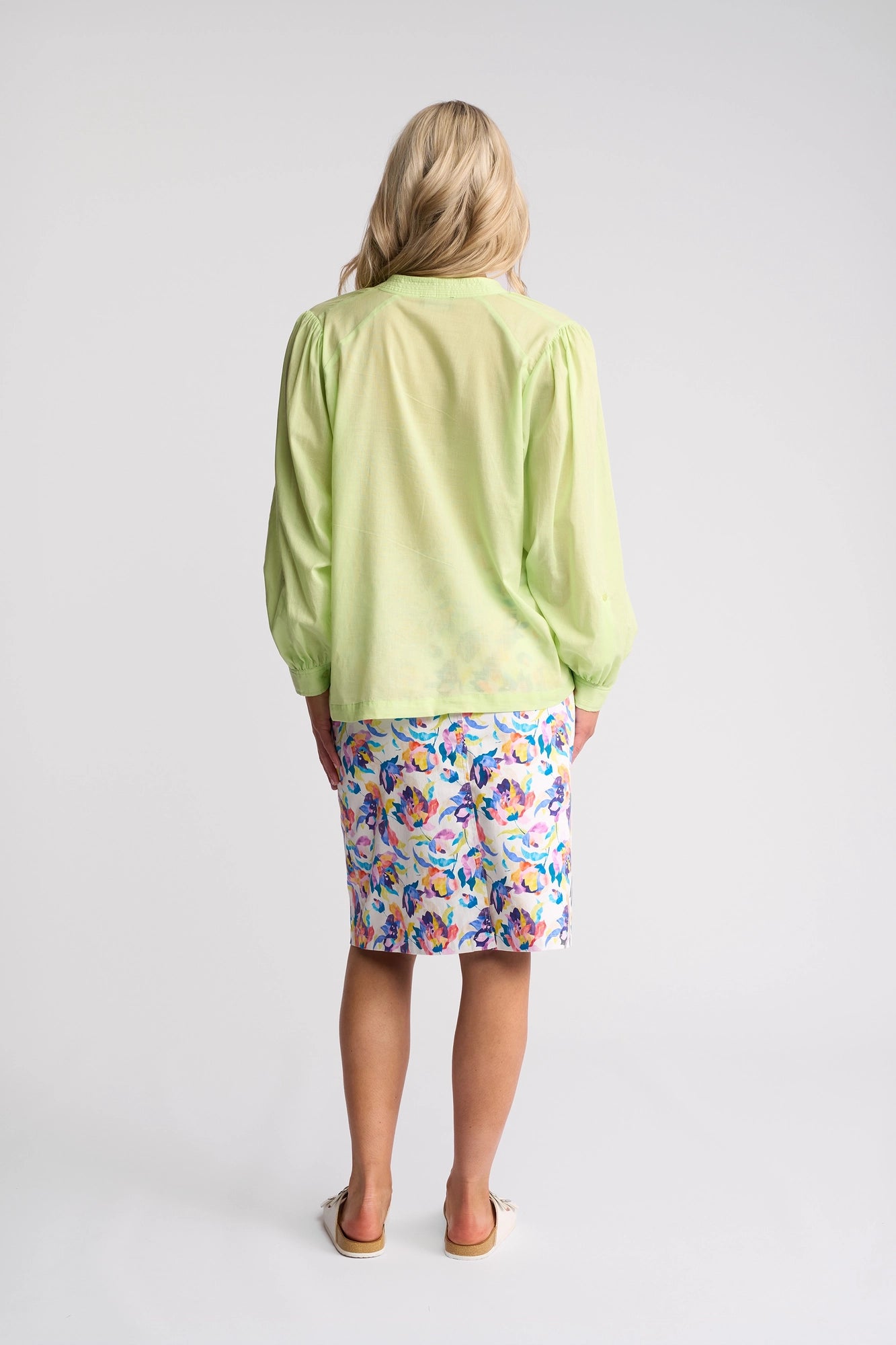 A woman with blonde hair stands facing away against a plain white background, wearing the Vassalli Lily Shirt in Citrus—a lightweight cotton shirt—paired with a colorful floral skirt.