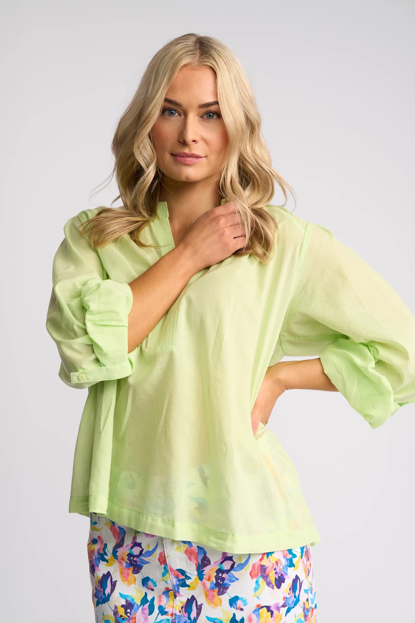 A woman with long blonde hair wears the Vassalli Lily Shirt in Citrus, a light green sheer blouse, styled with a floral skirt in blue, pink, and yellow hues, standing against a plain light background.