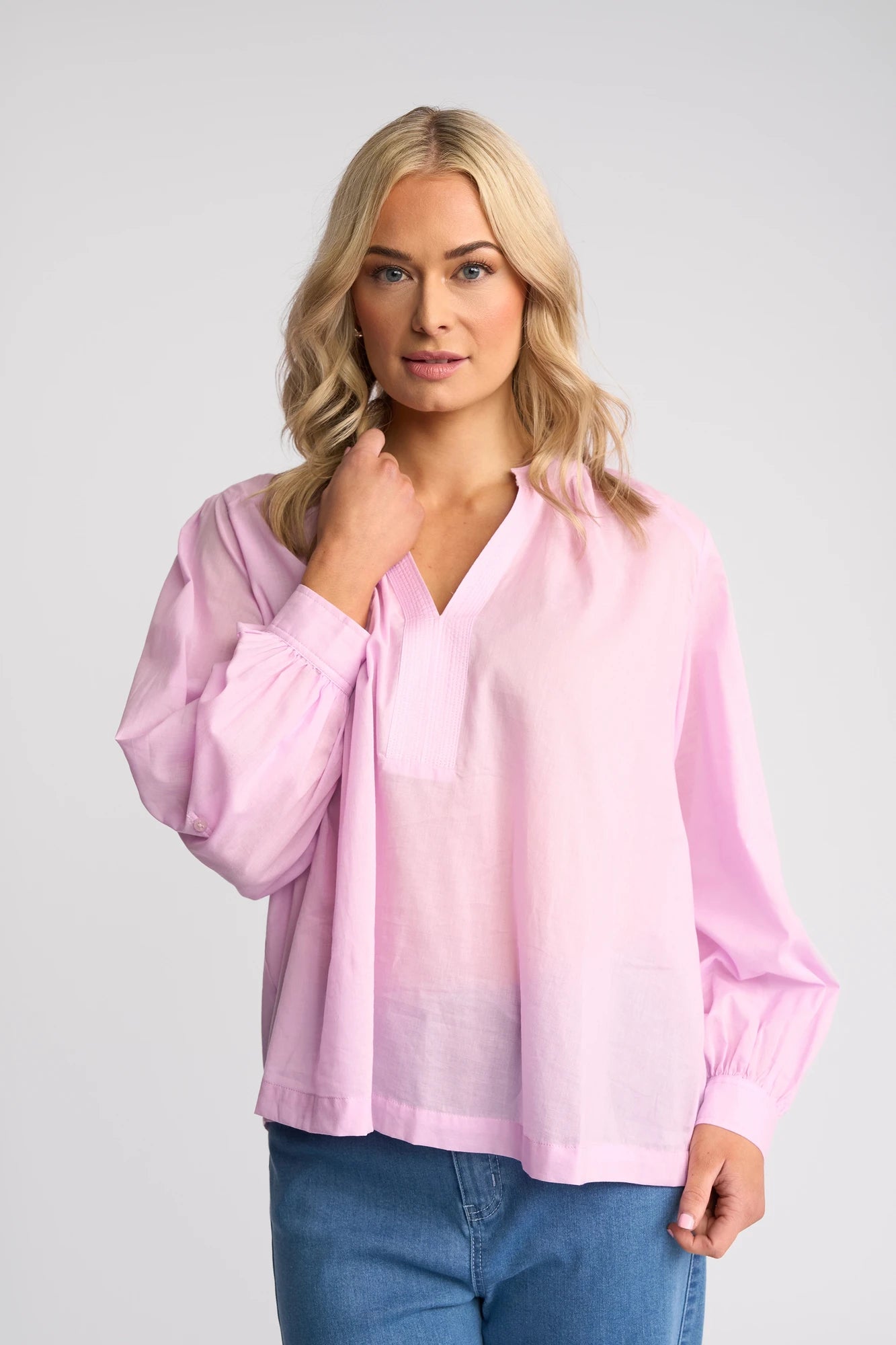 A woman with blonde hair wears the Vassalli Lily Shirt - Light Purple and blue jeans, standing against a plain gray background while looking at the camera.