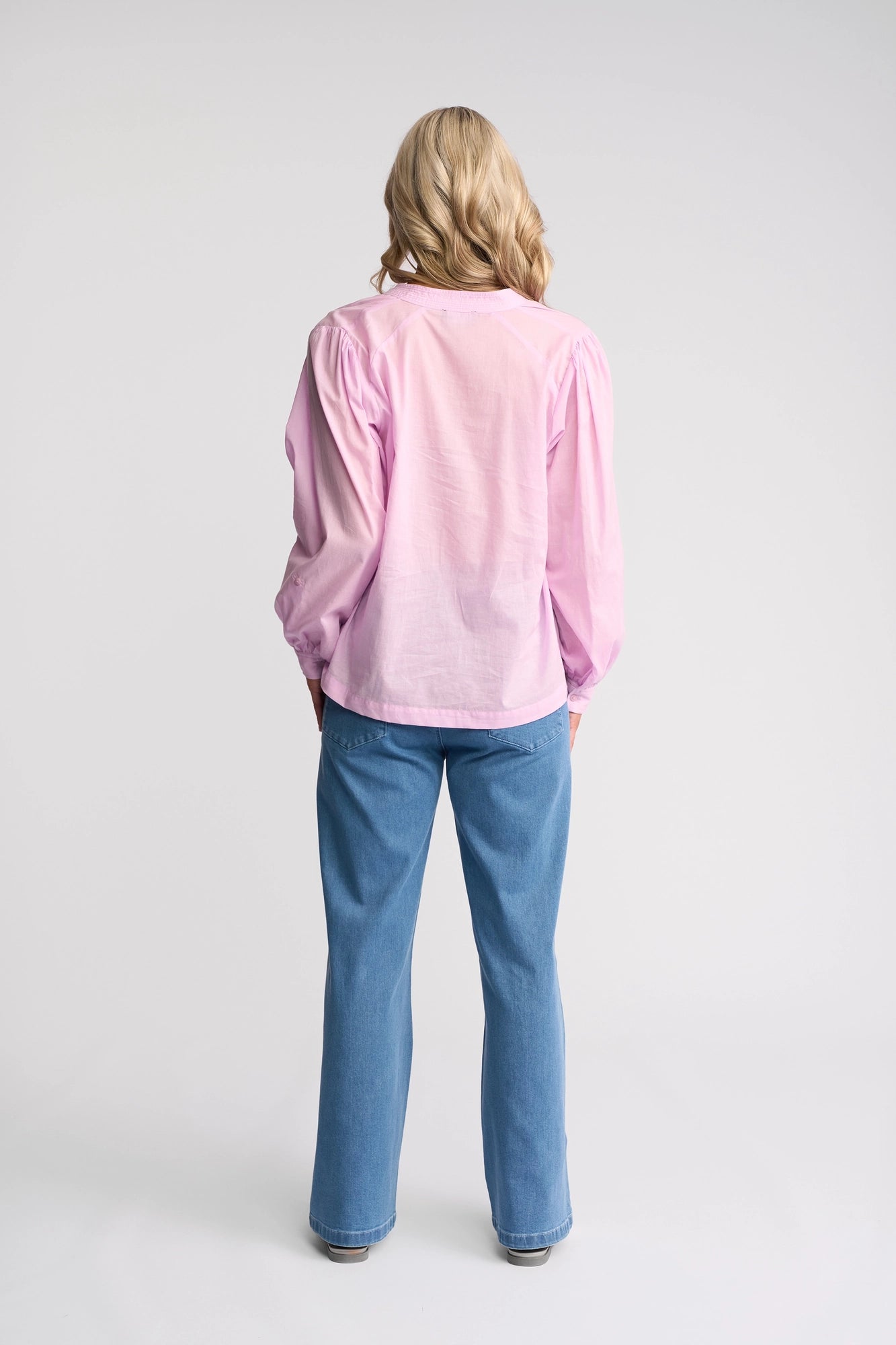 A person with long blonde hair stands facing away, wearing the Vassalli Lily Shirt in light purple and blue jeans, in a well-lit studio with a plain white background.