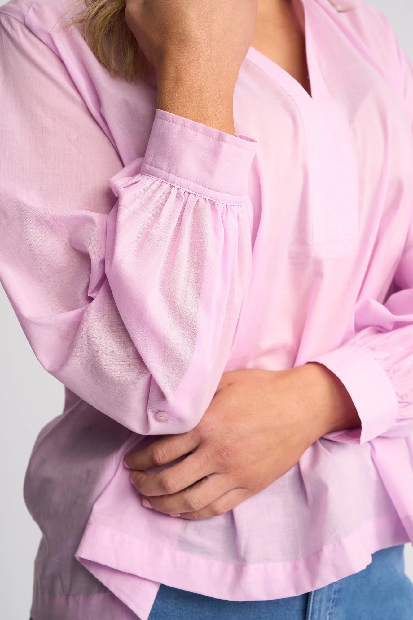 A person wearing a light purple, long-sleeved Vassalli Lily Shirt and blue jeans, with one hand resting on their stomach and the other hand touching their face. Their face is not fully visible.
