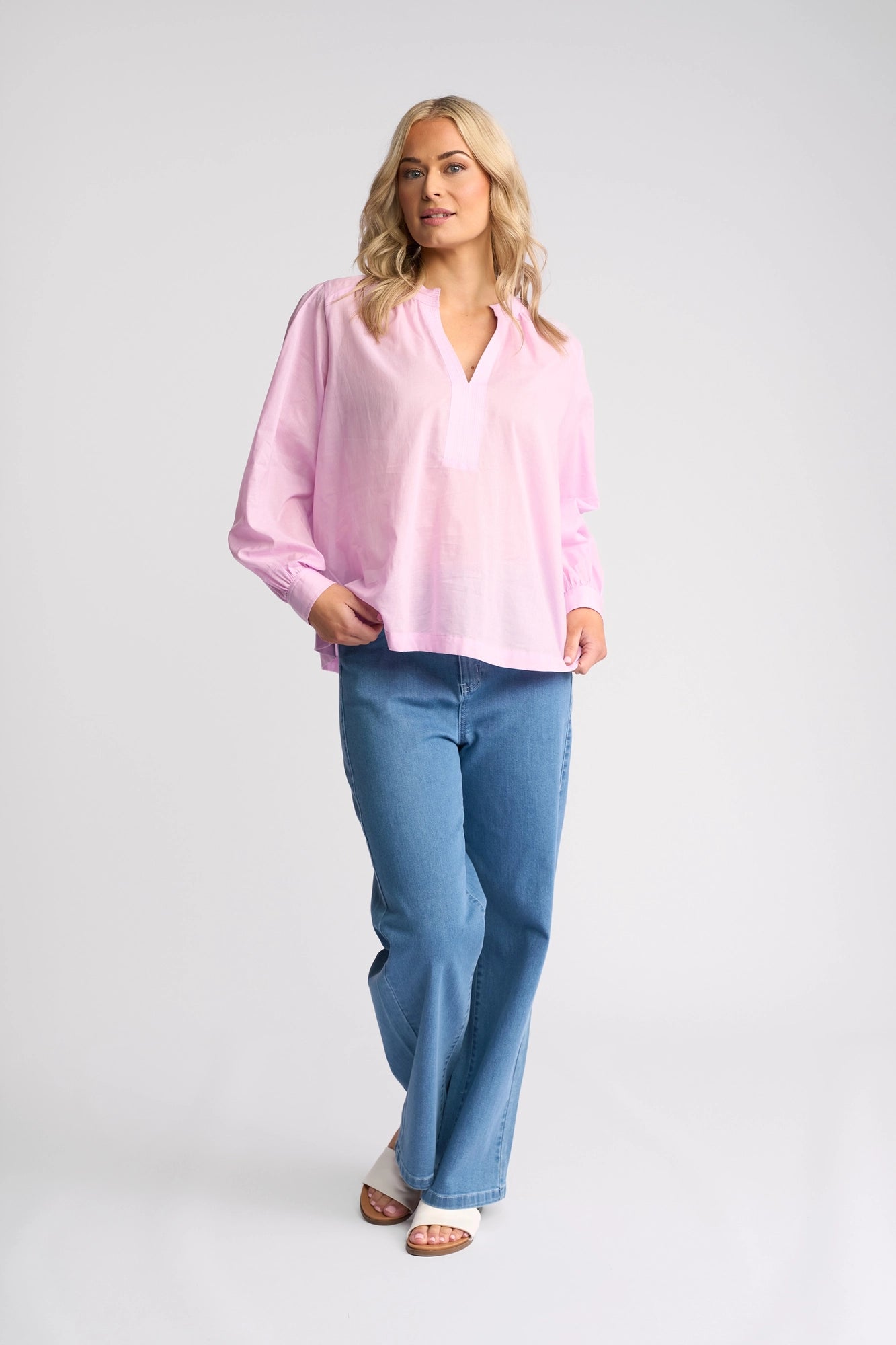 A woman stands against a white background wearing the Vassalli Lily Shirt in light purple, paired with blue jeans and white open-toe shoes. She has blonde hair, gazes slightly to the side, and has one hand in her pocket.