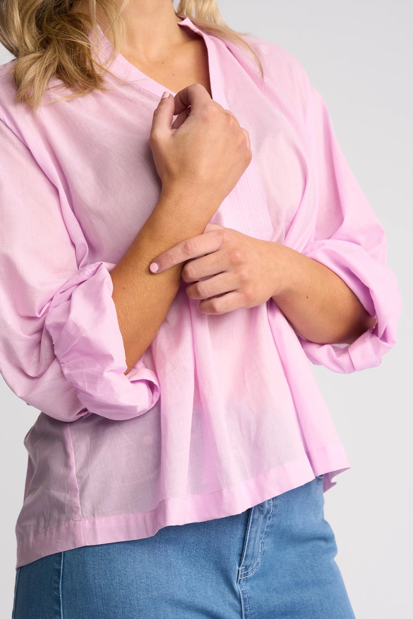 A person wearing a light purple Vassalli Lily Shirt and blue jeans touches their left forearm with their right hand. Only the upper body is visible; the face is not shown.