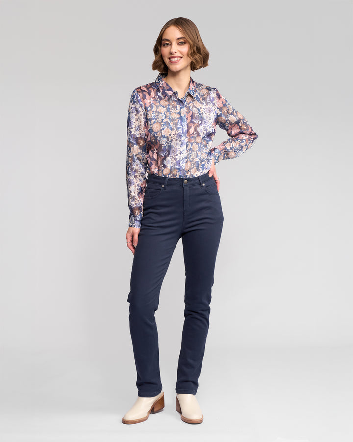 A woman smiles against a light gray background, wearing a long-sleeve floral blouse, Vassalli’s Column Pant/Jean in Carbon for a perfect fit, and off-white shoes. One hand rests on her hip while the other is by her side.