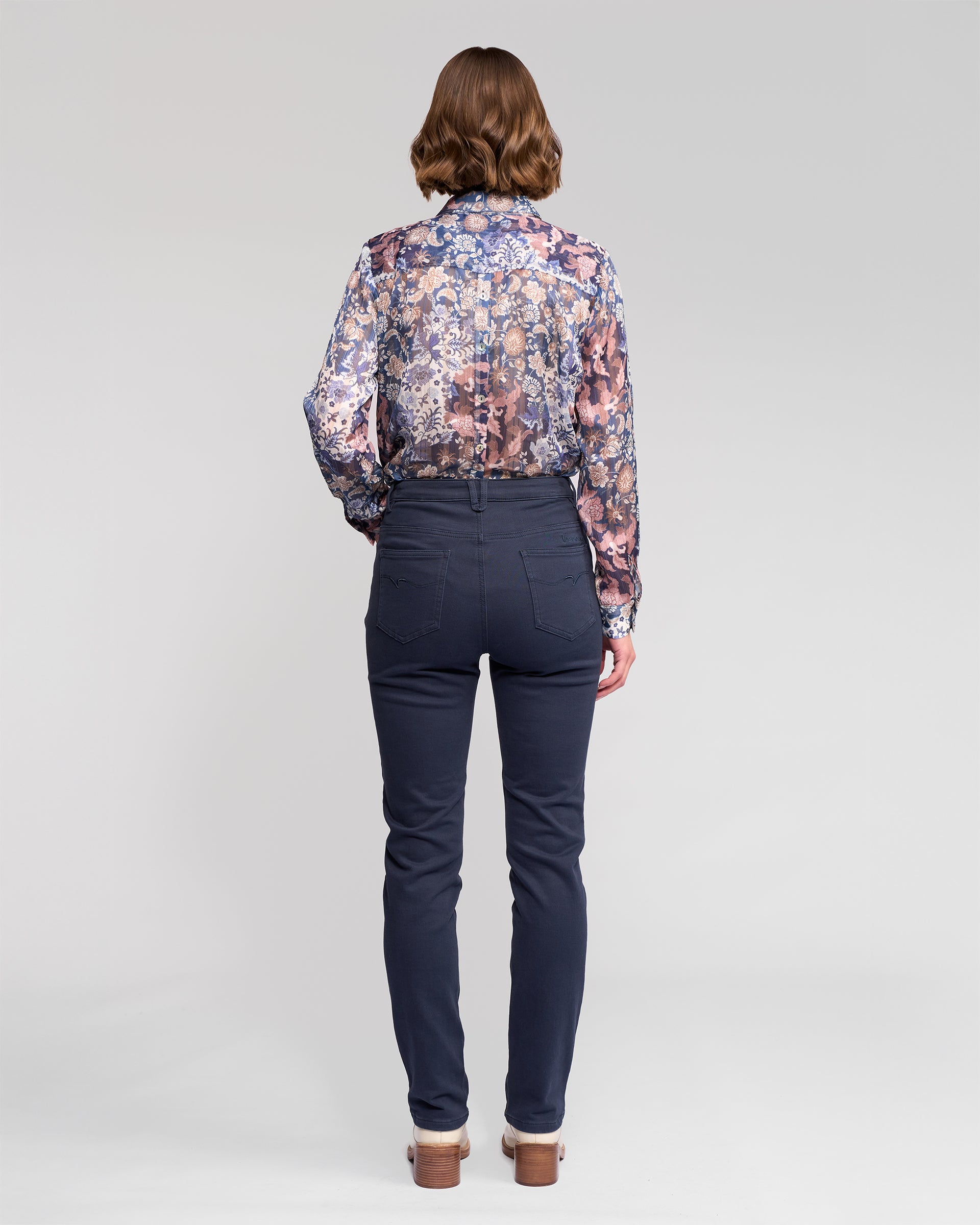 A woman with shoulder-length brown hair wears a sheer, long-sleeved floral blouse and Vassalli Column Pant/Jean in Carbon. Seen from behind, she stands against a plain light gray background, showcasing the jeans’ flattering fit and back pocket detail.