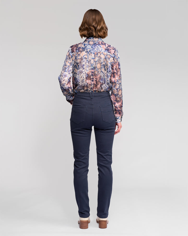 A woman with shoulder-length brown hair wears a sheer, long-sleeved floral blouse and Vassalli Column Pant/Jean in Carbon. Seen from behind, she stands against a plain light gray background, showcasing the jeans’ flattering fit and back pocket detail.