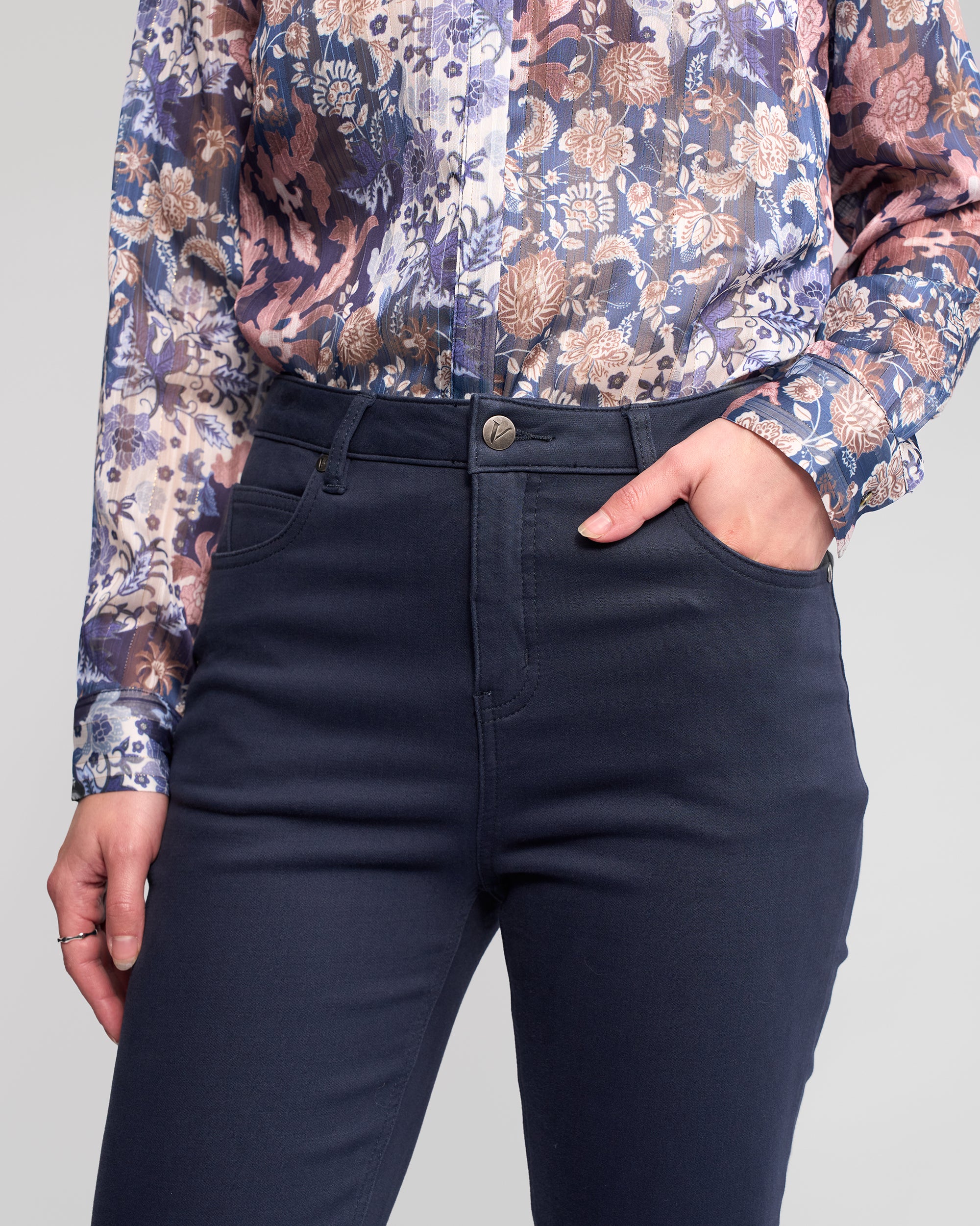 Wearing Vassalli's Column Pant/Jean in Carbon and a long-sleeve floral blouse in purple, blue, and beige, a person stands with one hand in their pocket for the perfect fit.