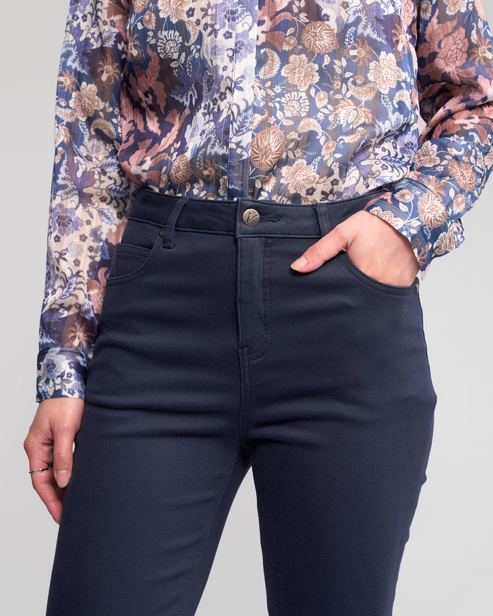 Wearing Vassalli's Column Pant/Jean in Carbon and a long-sleeve floral blouse in purple, blue, and beige, a person stands with one hand in their pocket for the perfect fit.