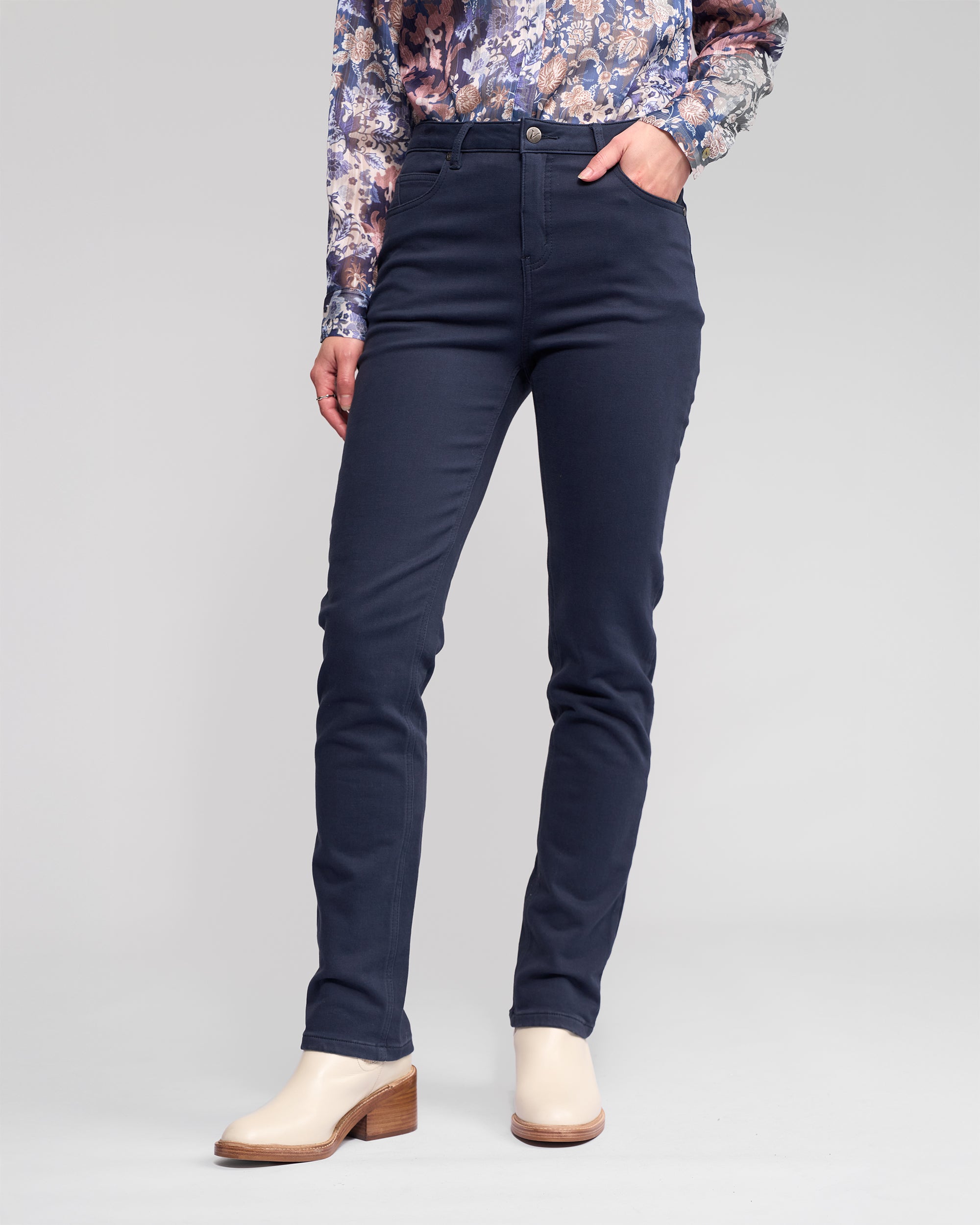 Someone wearing Vassalli's Column Pant/Jean in Carbon, paired with a floral long-sleeve shirt and light-toned heeled ankle boots, stands with one hand in their pocket against a plain background.