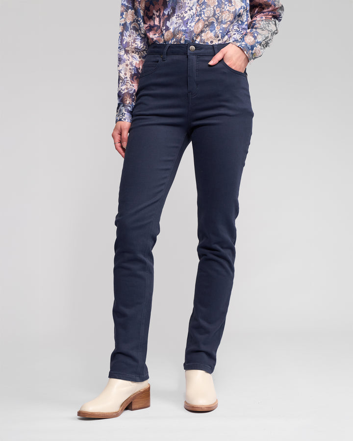 Someone wearing Vassalli's Column Pant/Jean in Carbon, paired with a floral long-sleeve shirt and light-toned heeled ankle boots, stands with one hand in their pocket against a plain background.