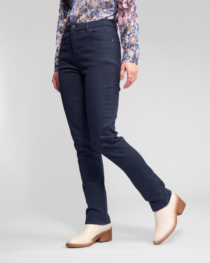 A person wearing Vassalli's Column Pant/Jean in Carbon, paired with a long-sleeved floral blouse and light heeled ankle boots, stands against a plain light background for the perfect fit.