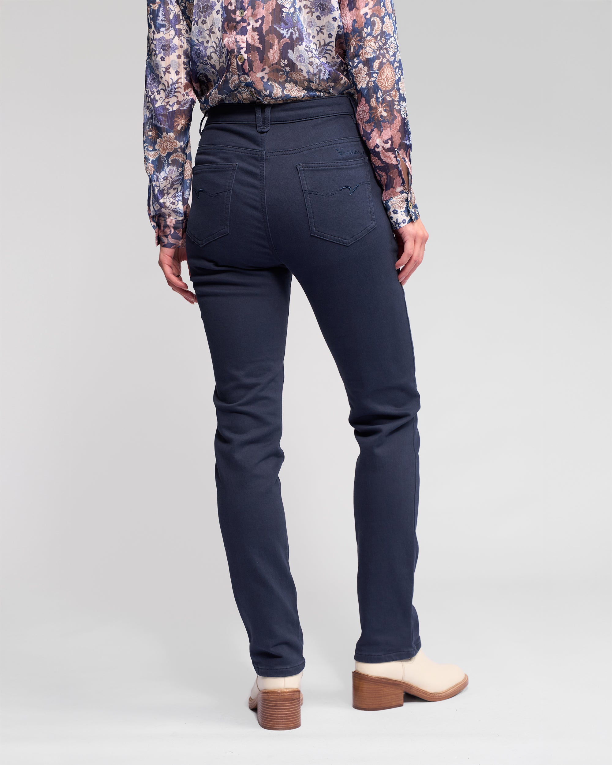 A person stands facing away, wearing Vassalli Column Pant/Jean in Carbon, a floral long-sleeve blouse, and beige heeled shoes against a plain light background.
