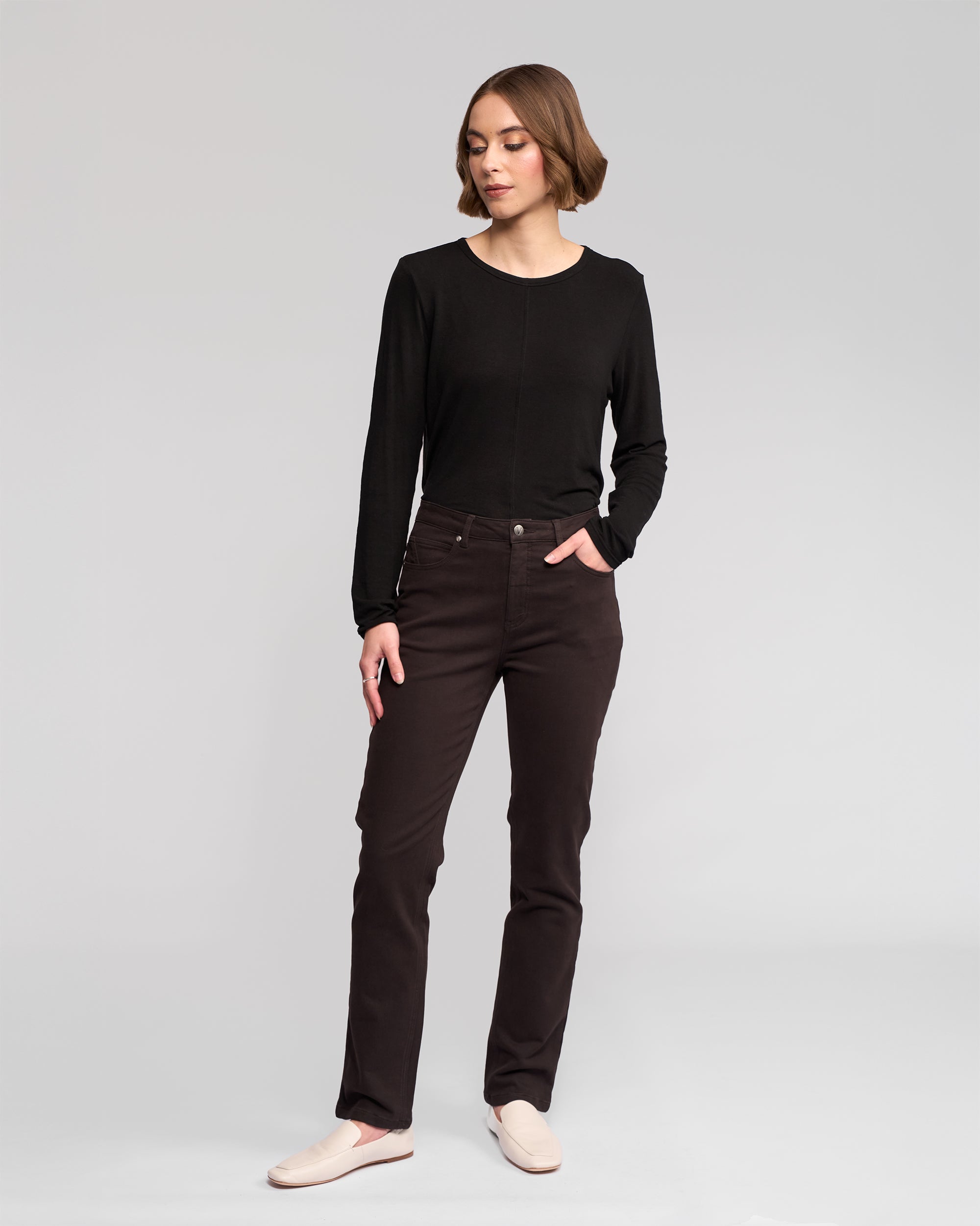 A woman stands against a plain light gray background, wearing a black long-sleeve top, Vassalli Column Pant/Jean in Chocolate, and light-colored slip-on shoes. She looks slightly to the side with one hand in her pocket.