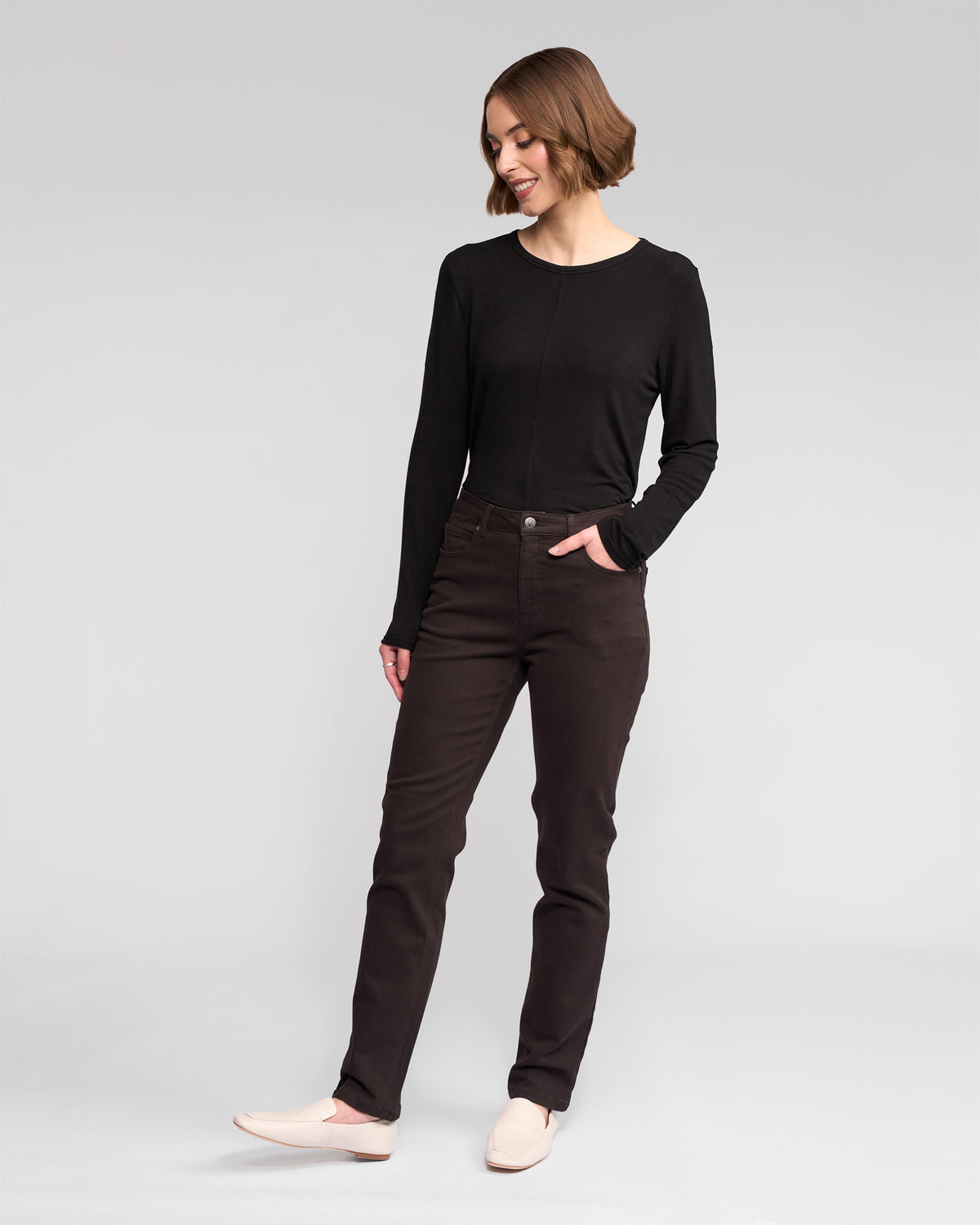 A woman with short brown hair wears a Vassalli Column Pant/Jean in chocolate, a long-sleeve black top, and light slip-on shoes. She stands with one hand in her pocket, looking down and smiling slightly against a plain background.