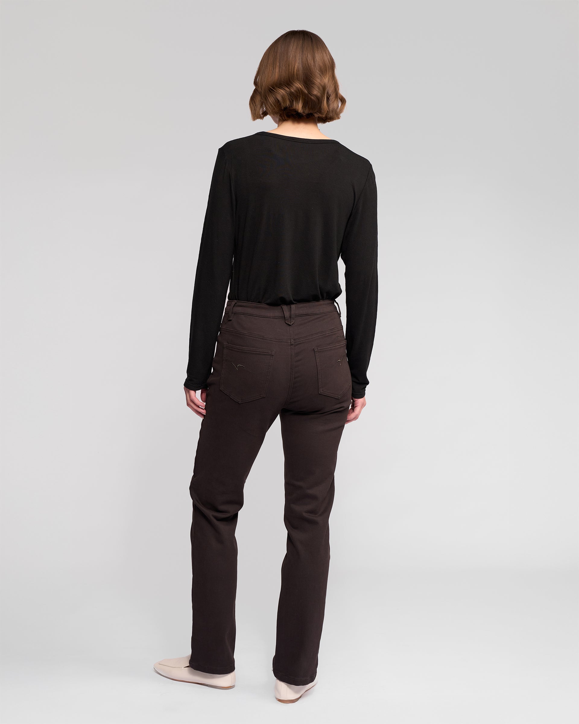 A person with short brown hair, wearing a long-sleeve black top and Vassalli Column Pant/Jean in Chocolate, stands facing away from the camera against a plain light gray background.