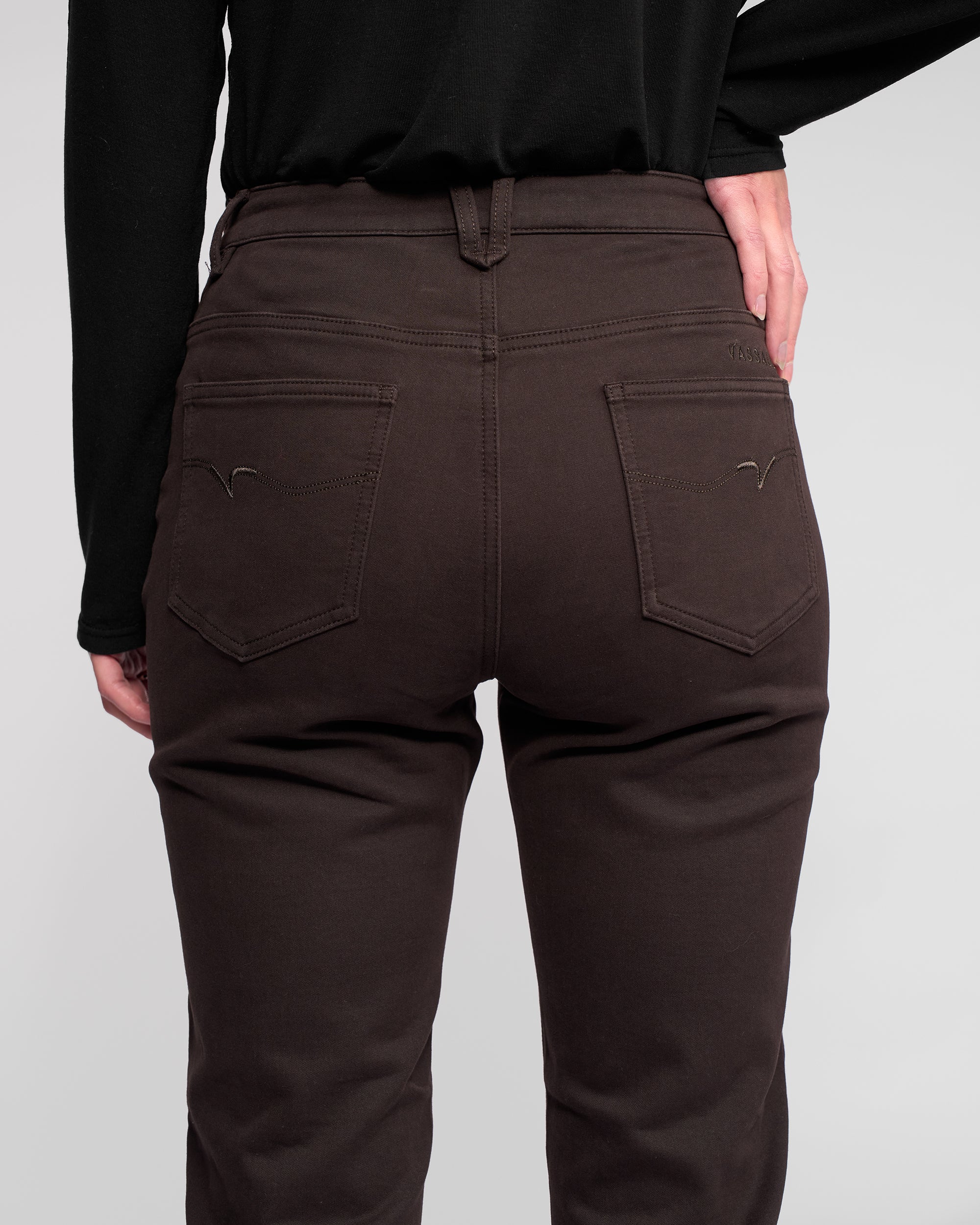 A person is shown from the back wearing Vassalli Column Pant/Jean in Chocolate and a black long-sleeve shirt, with one hand on their right hip against a plain light background.