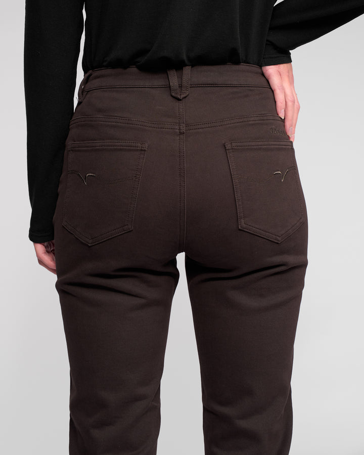 A person is shown from the back wearing Vassalli Column Pant/Jean in Chocolate and a black long-sleeve shirt, with one hand on their right hip against a plain light background.
