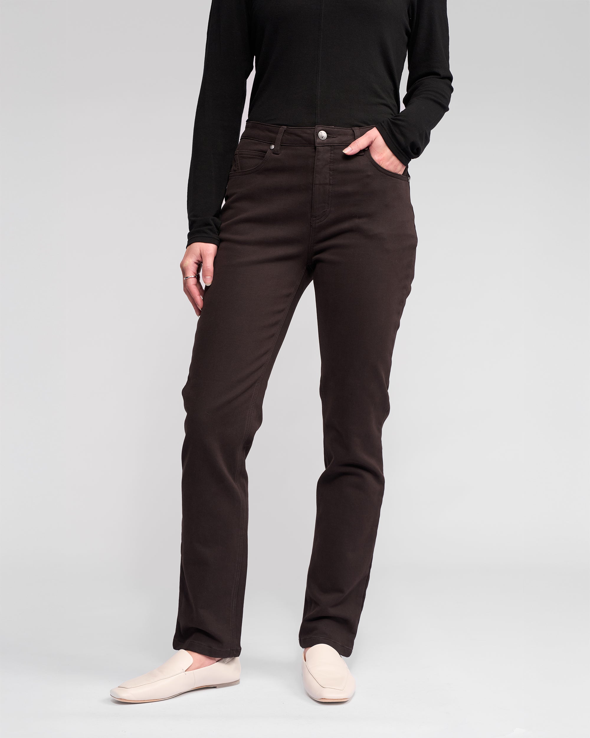 A person stands against a plain light background with one hand in their pocket, wearing chocolate Column Pant/Jean by Vassalli, a black long-sleeve top, and beige slip-on shoes. Their face is not visible.