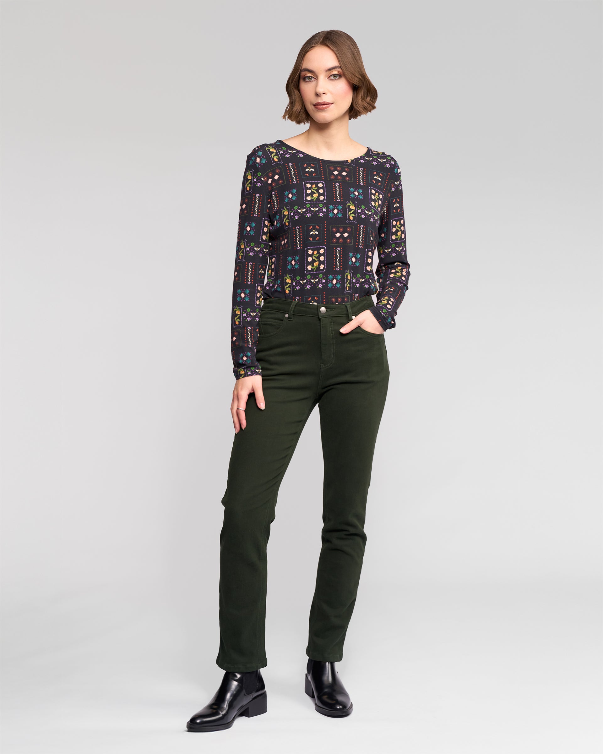 A woman with short brown hair stands against a light background, wearing a patterned dark long-sleeve top and Vassalli Column Pant/Jean in Forest, paired with black ankle boots, looking at the camera with a relaxed posture.